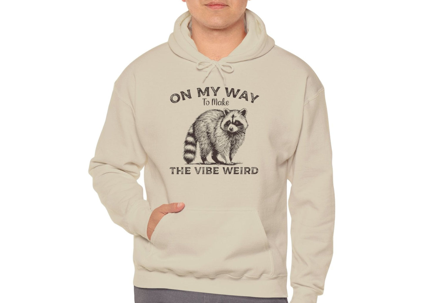 Model wearing a light blue hoodie featuring the text "On My Way to Make the Vibe Weird" and a cartoon illustration of a raccoon holding a stick. Part of the Vivid Divergence Sensory Friendly Unisex Hoodie Range.