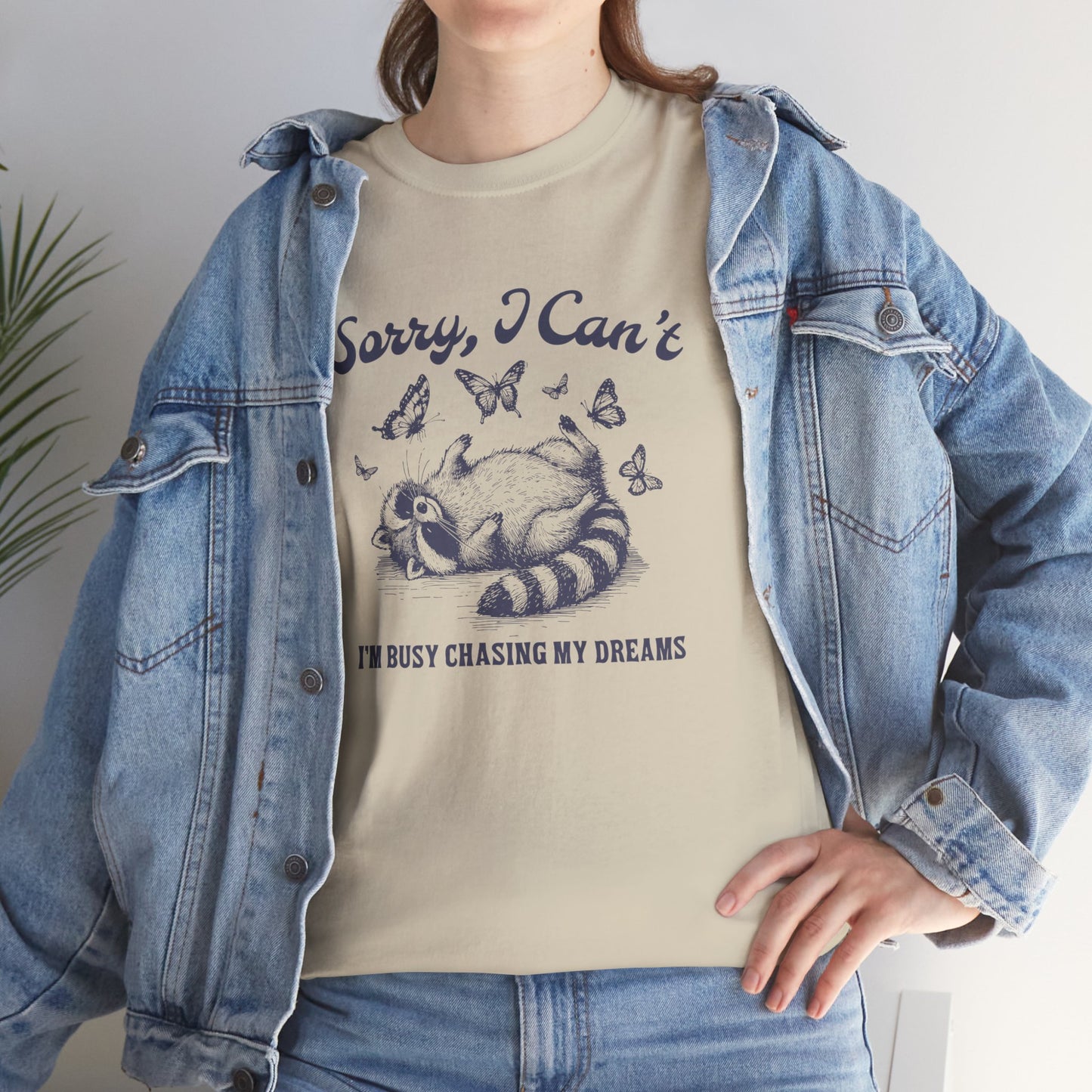 Model wearing a beige T-shirt with "Sorry I Can't, I'm Busy Chasing My Dreams" and a playful sleeping raccoon graphic in black text. Part of the Vivid Divergence Sensory Friendly Unisex Tees Range.