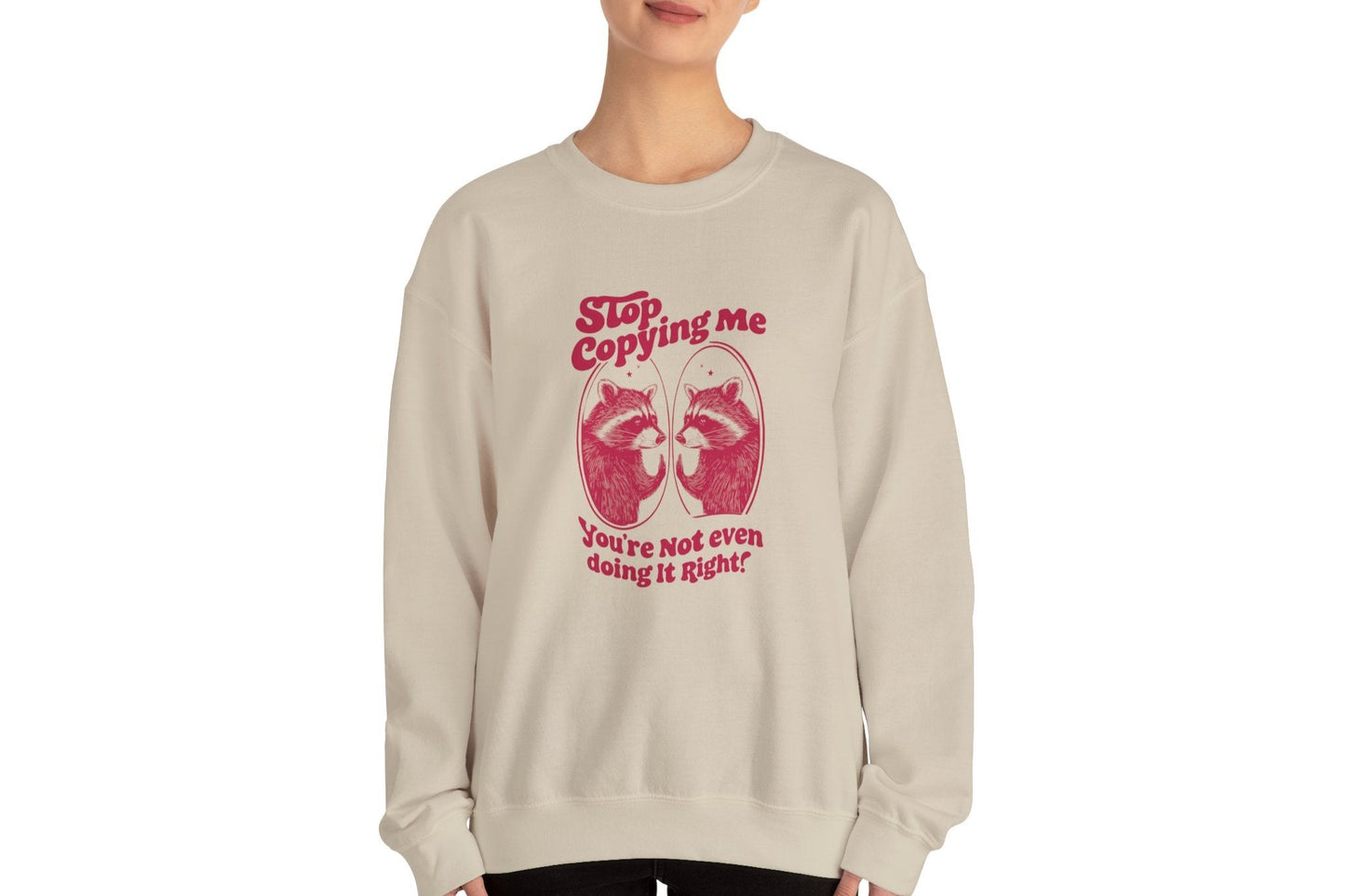 Model wearing a beige sweater jumper with the text "Stop Copying Me, You’re Not Even Doing It Right," featuring a cartoon raccoon looking in a mirror. Part of the Vivid Divergence Sensory Friendly Unisex Sweater Jumper Range.