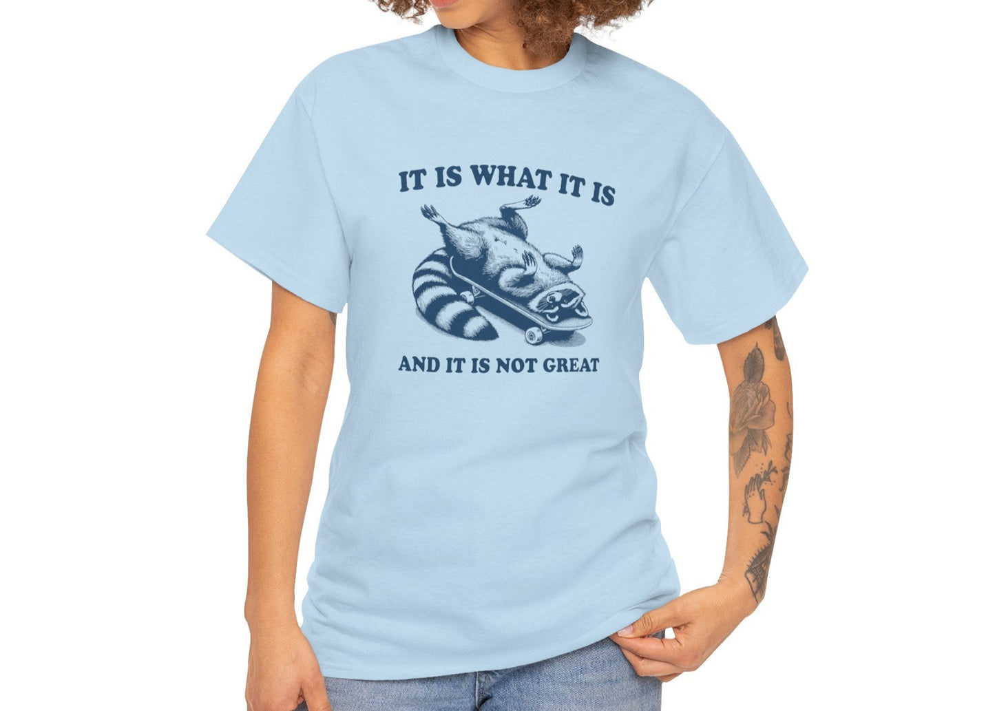 Female model wearing a light blue T-shirt with a raccoon on a skateboard and "It Is What It Is... And It's Not Great" text. Part of the Vivid Divergence Sensory Friendly Unisex Tees Range.