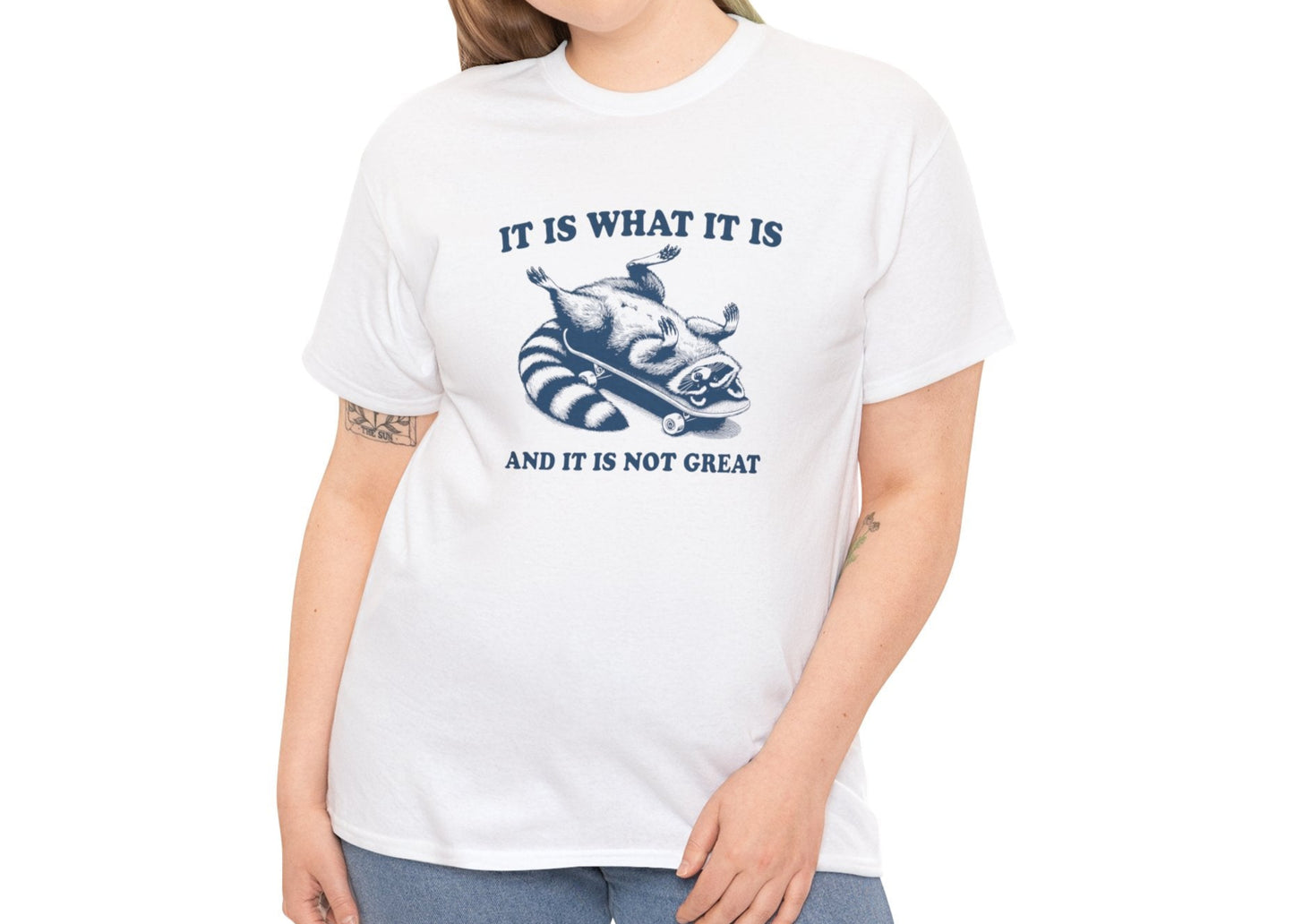 Female model wearing a white T-shirt with a blue raccoon on a skateboard and "It Is What It Is... And It's Not Great" text. Part of the Vivid Divergence Sensory Friendly Unisex Tees Range.