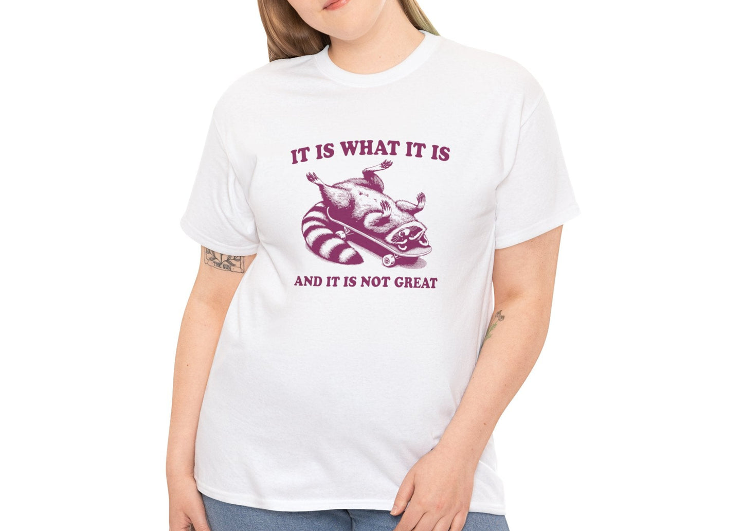 Female model wearing a white T-shirt featuring a raccoon on a skateboard with "It Is What It Is... And It's Not Great" text in pink. Part of the Vivid Divergence Sensory Friendly Unisex Tees Range.