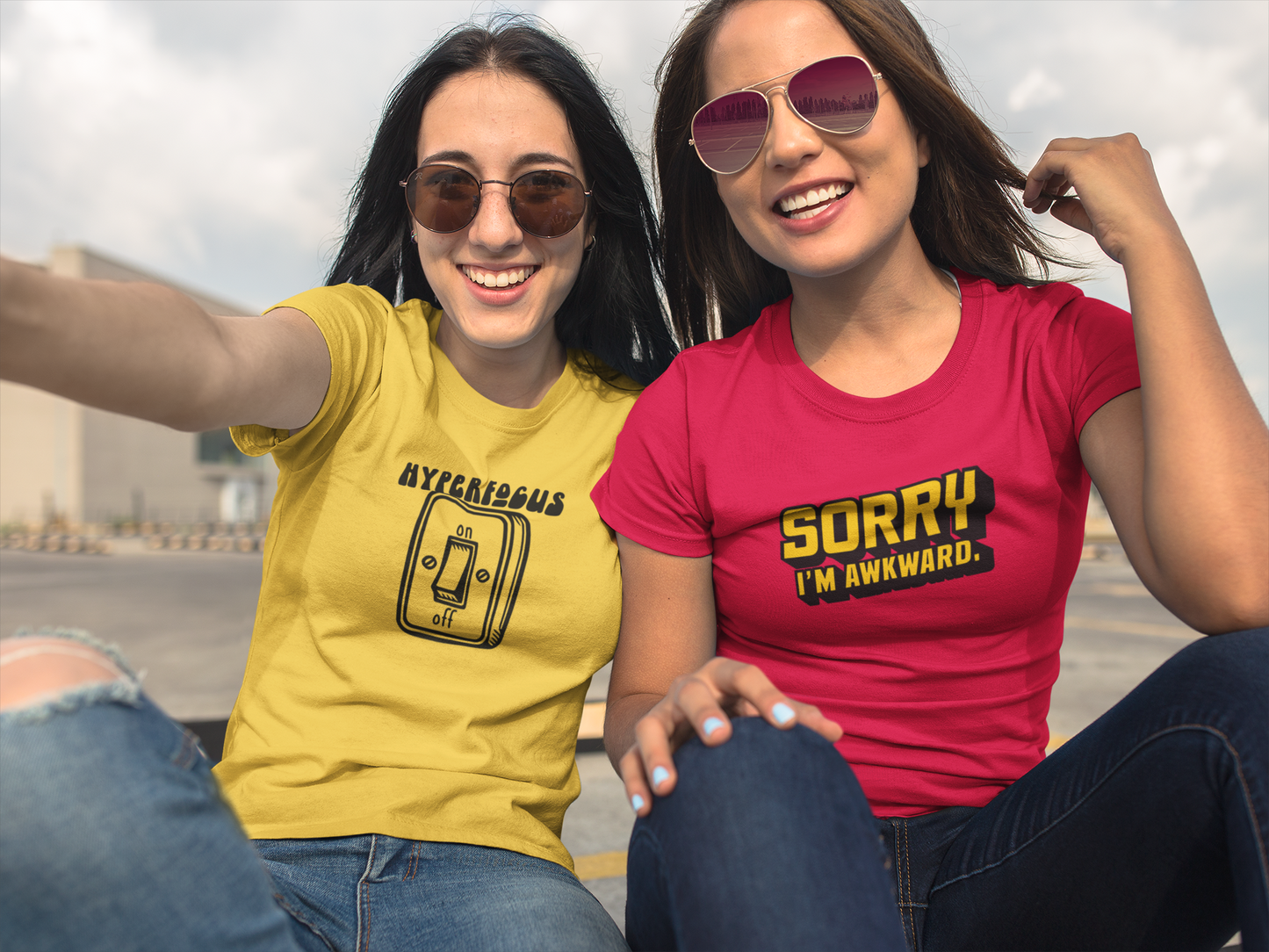 Two people sitting together, one wearing a red T-shirt and the other a yellow T-shirt, both featuring the text "Hyperfocus" with an on/off switch graphic design. Part of the Vivid Divergence Sensory Friendly Unisex Tees Range.
