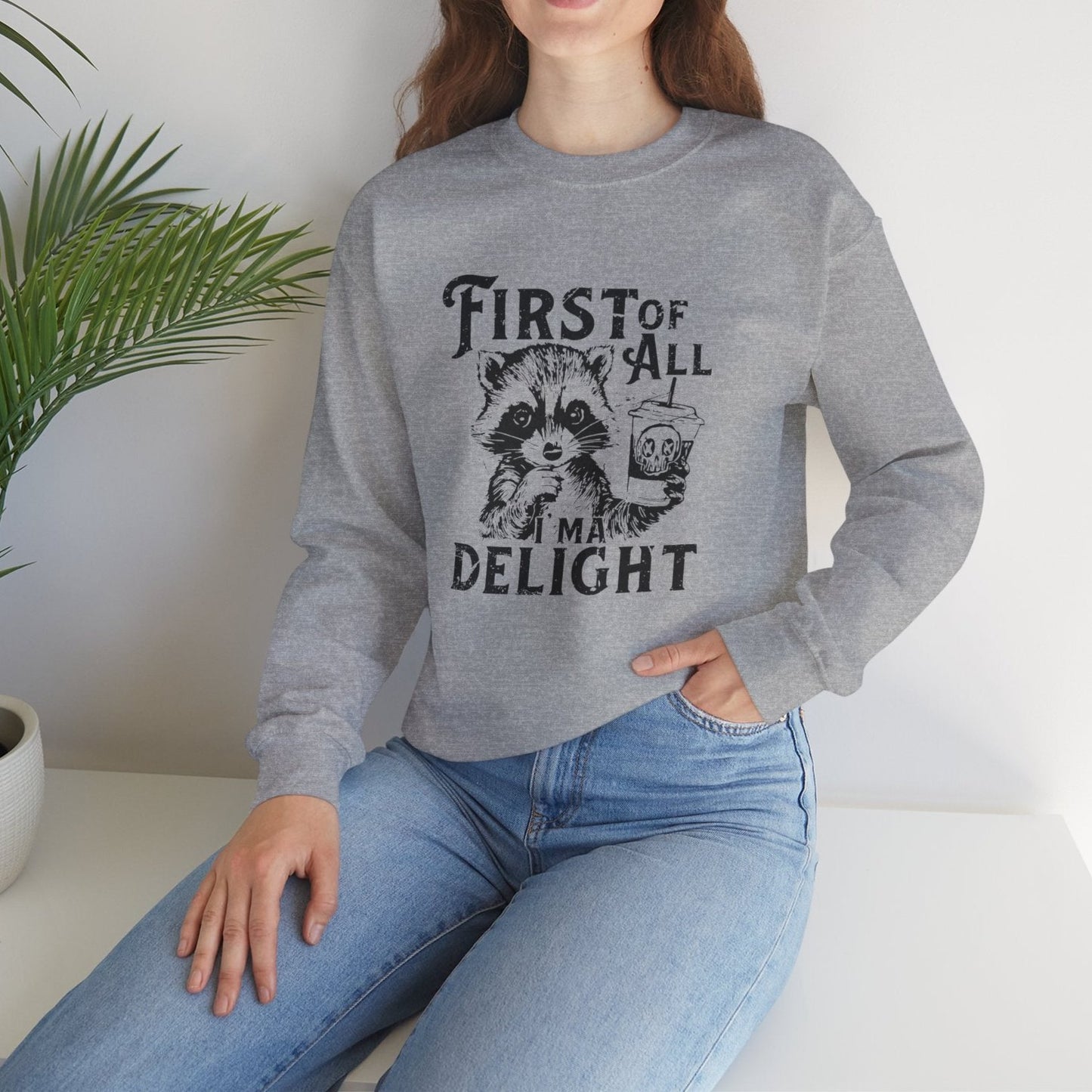 Model wearing a gray sweater jumper with the text "First of All, I’m a Delight" and a cartoon raccoon illustration. Part of the Vivid Divergence Sensory Friendly Unisex Sweater Jumper Range.