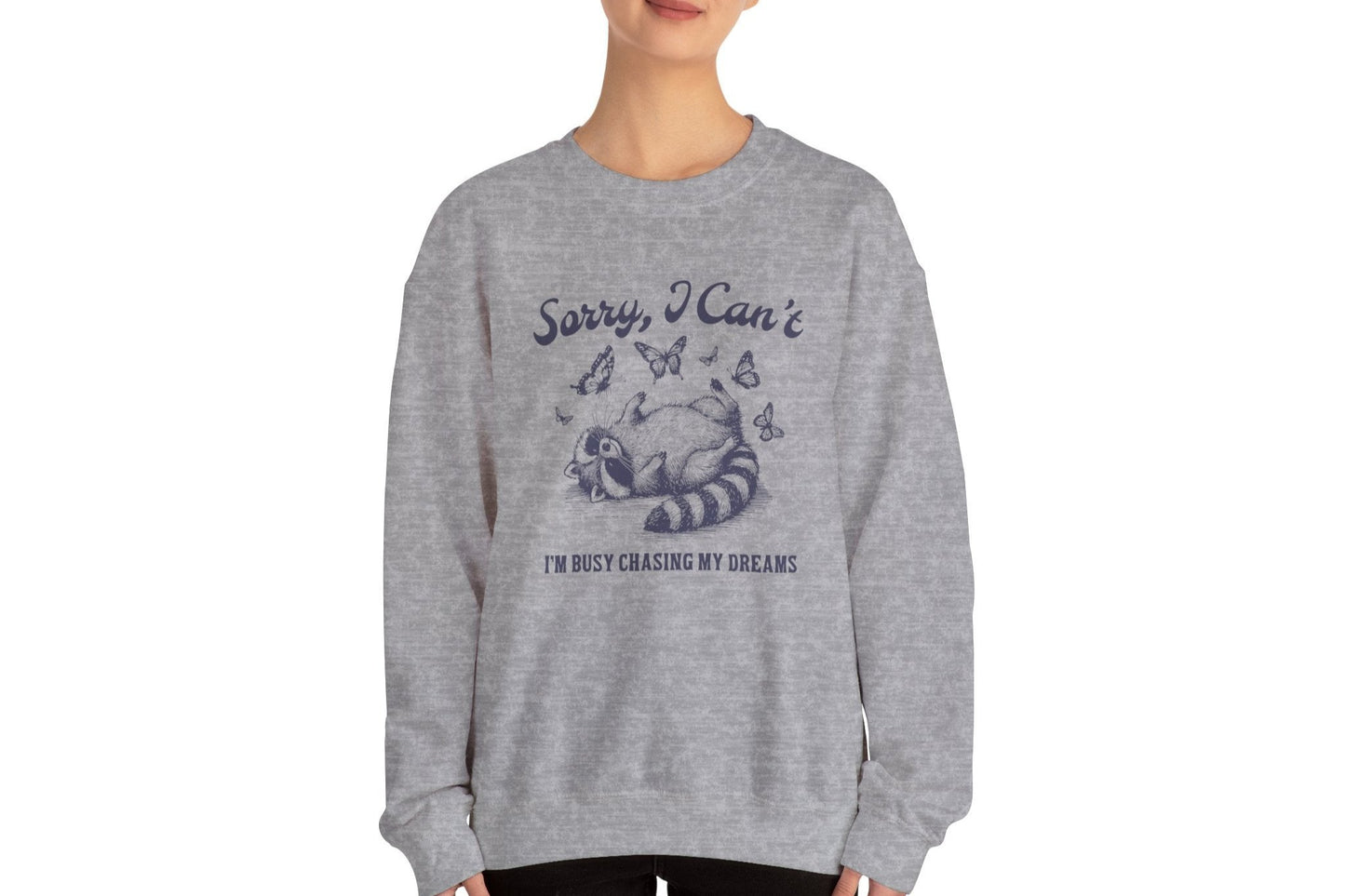 Model wearing a gray sweater jumper with the text "Sorry, I Can't. I'm Busy Chasing My Dreams" and a cartoon raccoon illustration sleeping on a crescent moon. Part of the Vivid Divergence Sensory Friendly Unisex Sweater Jumper Range.