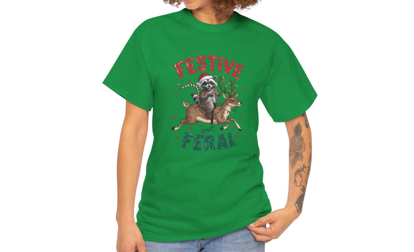 Model wearing a green T-shirt featuring a festive raccoon holding Christmas lights and the text "Festive but Feral," paired with casual jeans. Part of the Vivid Divergence Sensory Friendly Unisex Tees Range.