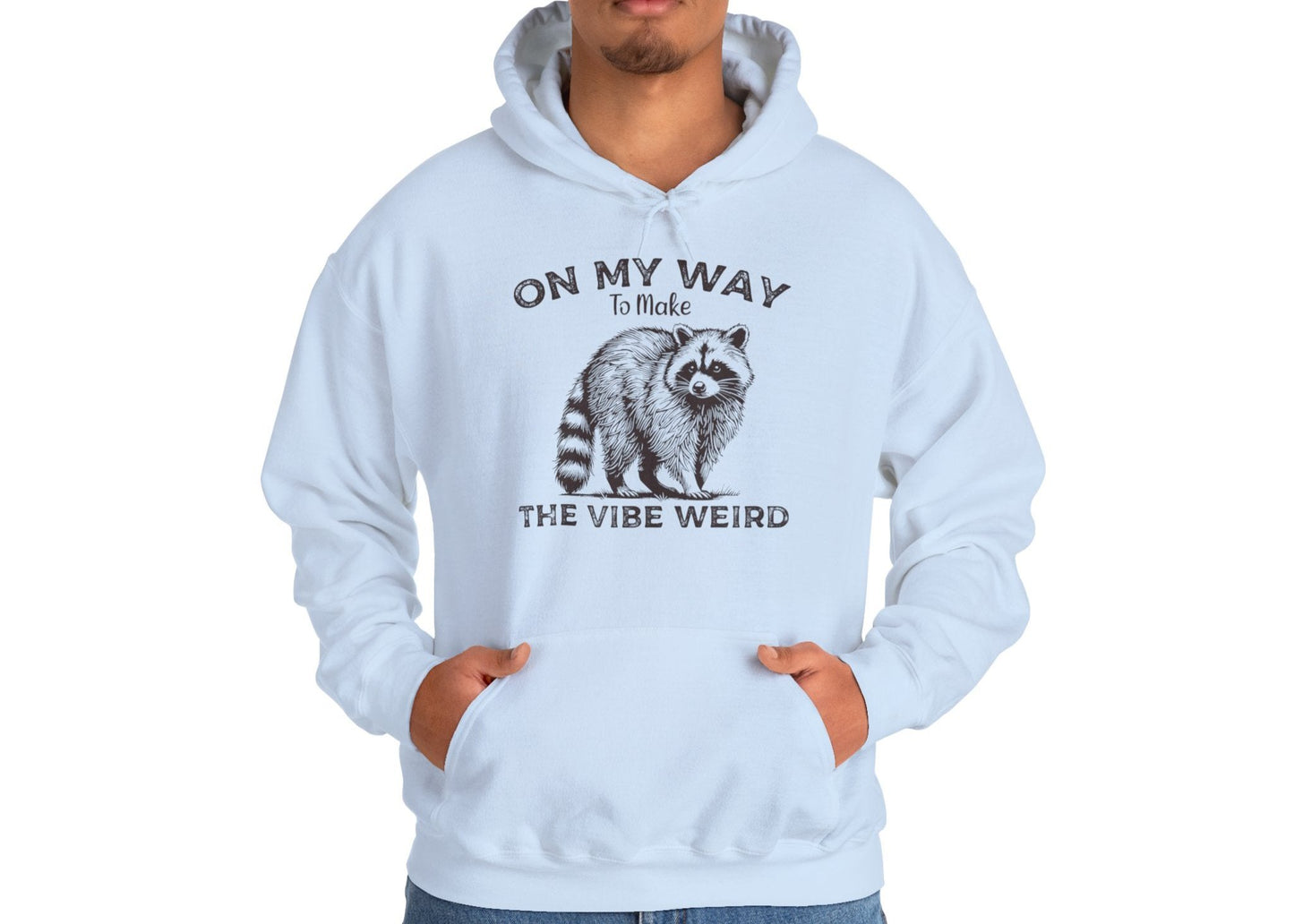 Model wearing a light blue hoodie featuring the text "On My Way to Make the Vibe Weird" and a cartoon illustration of a raccoon holding a stick. Part of the Vivid Divergence Sensory Friendly Unisex Hoodie Range.