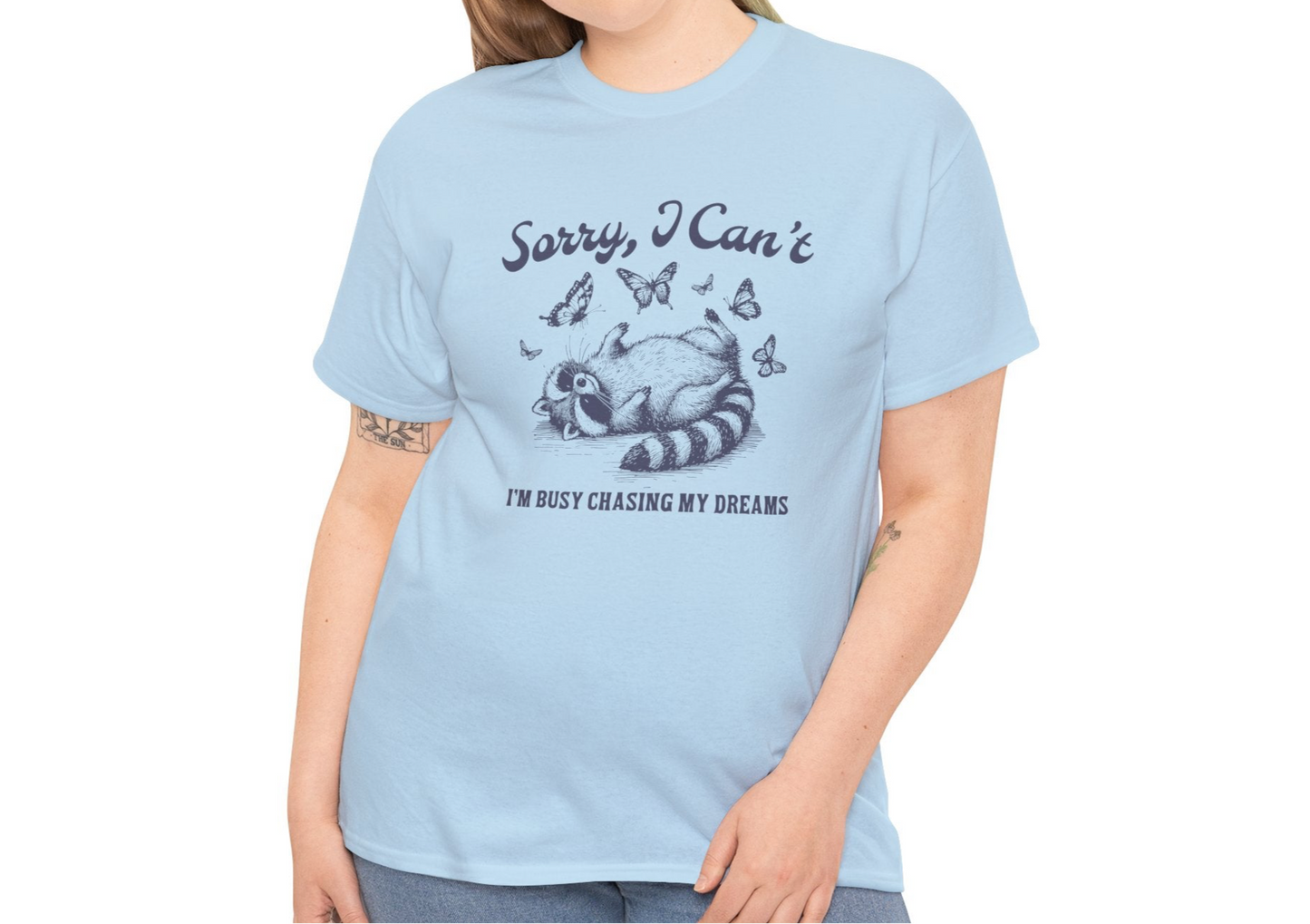 Model wearing a light blue T-shirt with "Sorry I Can't, I'm Busy Chasing My Dreams" and a playful sleeping raccoon graphic in black text. Part of the Vivid Divergence Sensory Friendly Unisex Tees Range.