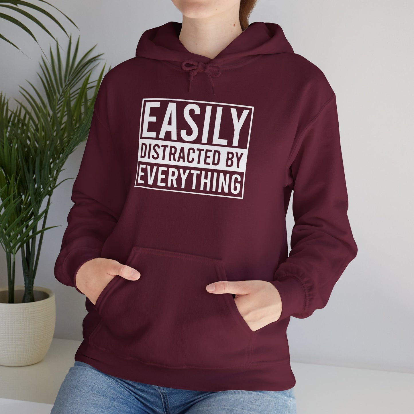 Model wearing a maroon hoodie with the text "Easily Distracted by Everything" in bold white lettering. Part of the Vivid Divergence Sensory Friendly Unisex Hoodie Range.