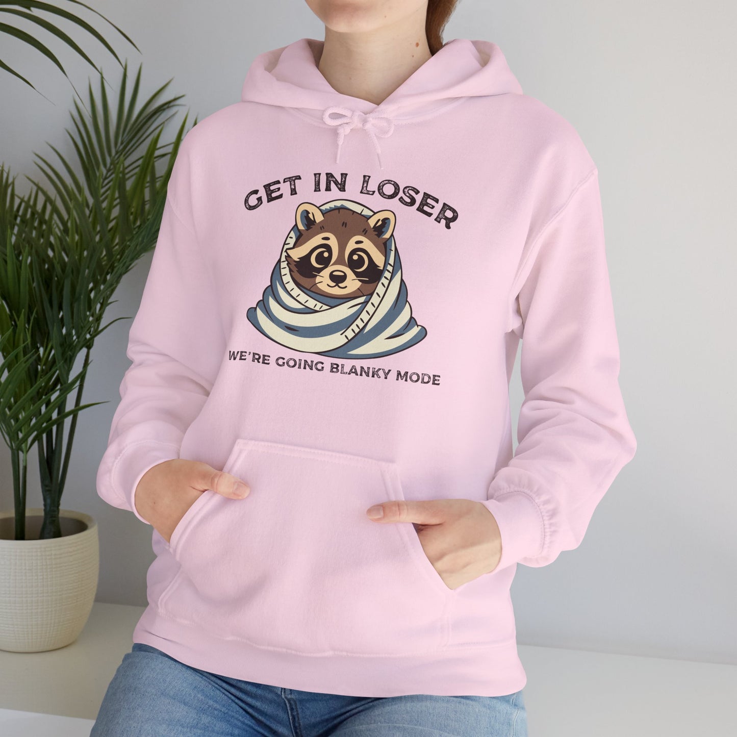 Model wearing a pink hoodie with a raccoon wrapped in a blanket and the text "Get in Loser, We're Going Blanky Mode," adjusting the hood. Part of the Vivid Divergence Sensory Friendly Unisex Hoodie Range.