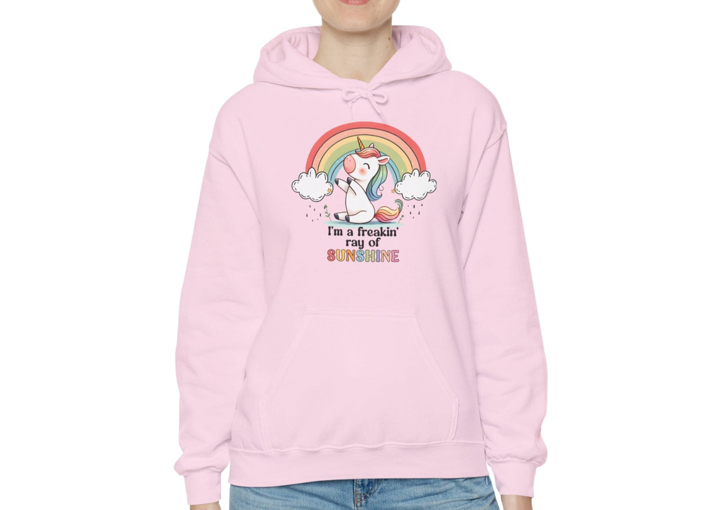 Model wearing a pink hoodie with a grumpy-looking unicorn under a rainbow and the text "I'm a Freakin' Ray of Sunshine," adjusting the hood. Part of the Vivid Divergence Sensory Friendly Unisex Hoodie Range.