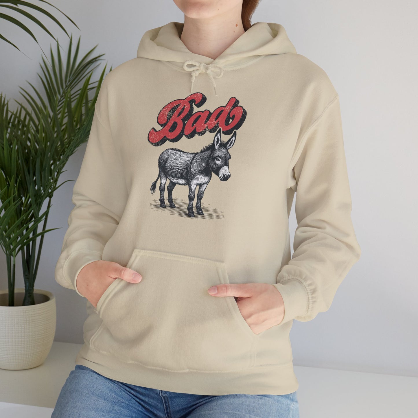 Model wearing a beige hoodie with the word "Bad" in bold red letters and a donkey illustration, standing with hands in pockets. Part of the Vivid Divergence Sensory Friendly Unisex Hoodie Range.