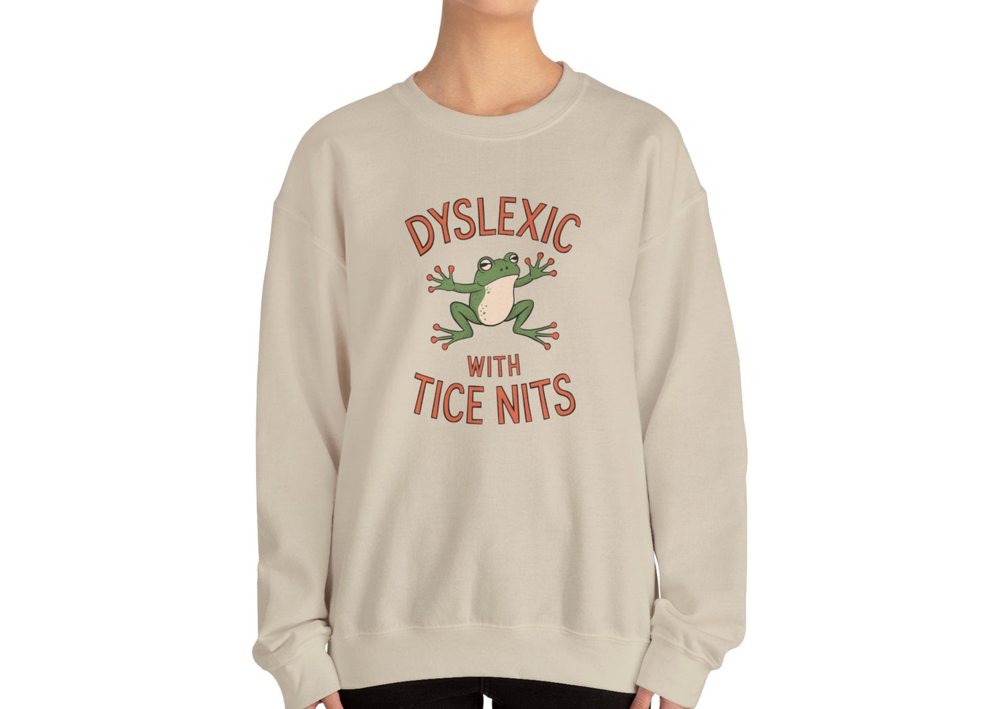 Model wearing a beige sweatshirt with a cartoon frog and the text "Dyslexic with Tice Nits," standing with hands at sides. Part of the Vivid Divergence Sensory Friendly Unisex Sweater Jumper Range.