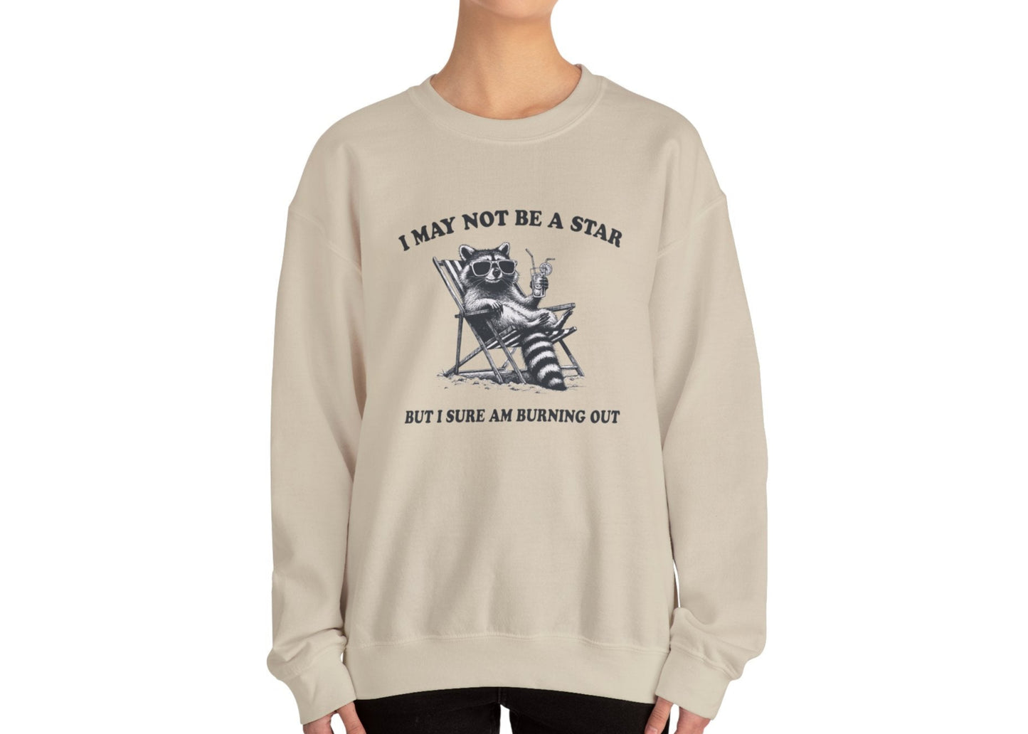 Model wearing a beige sweatshirt with a raccoon sitting in a trash can and the text "I May Not Be A Star, But I Sure Am Burning Out," standing with hands at sides. Part of the Vivid Divergence Sensory Friendly Unisex Sweater Jumper Range.