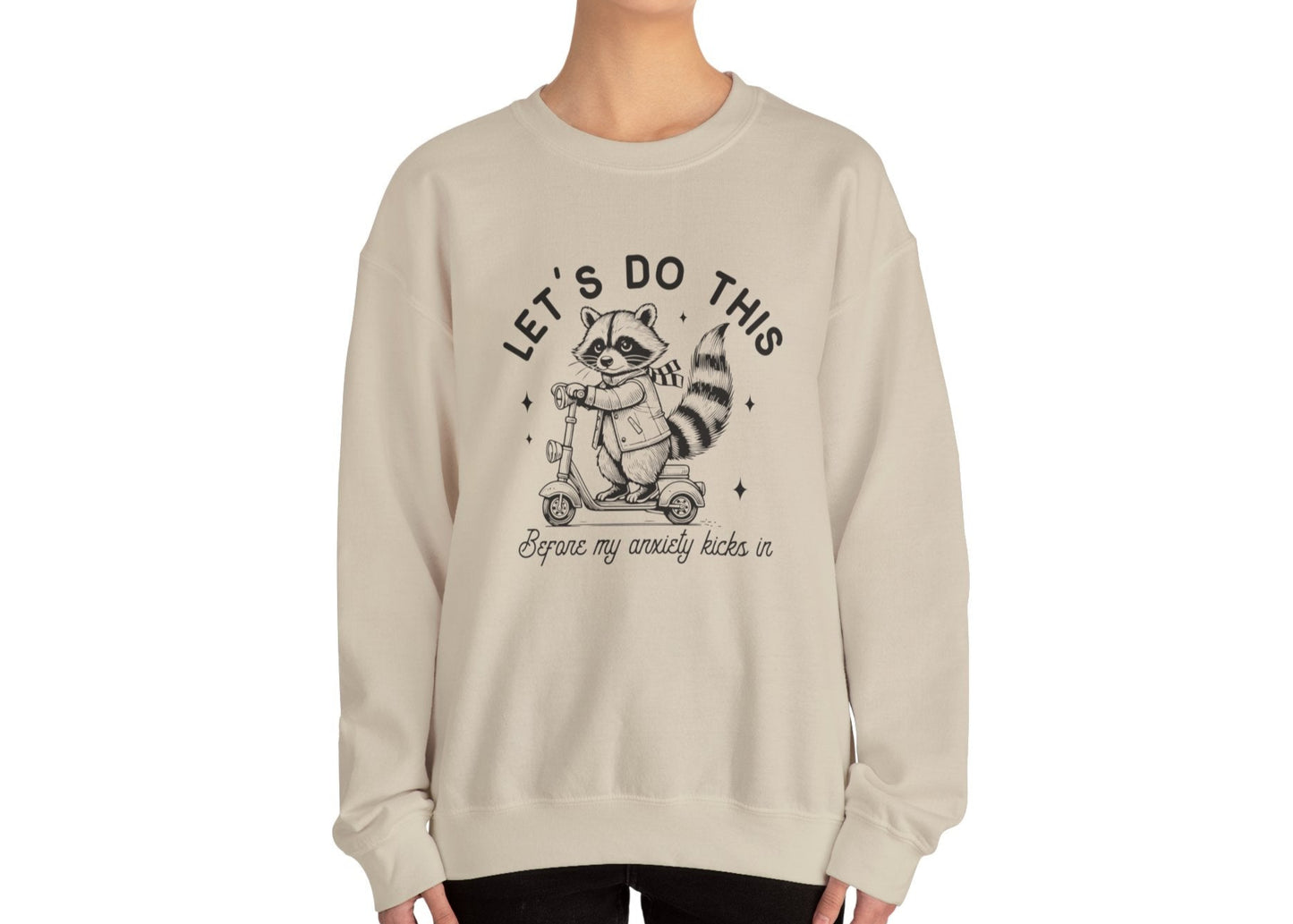 Model wearing a beige sweatshirt with a cartoon raccoon riding a scooter and the text "Let's Do This Before My Anxiety Kicks In," standing with hands at sides. Part of the Vivid Divergence Sensory Friendly Unisex Sweater Jumper Range.