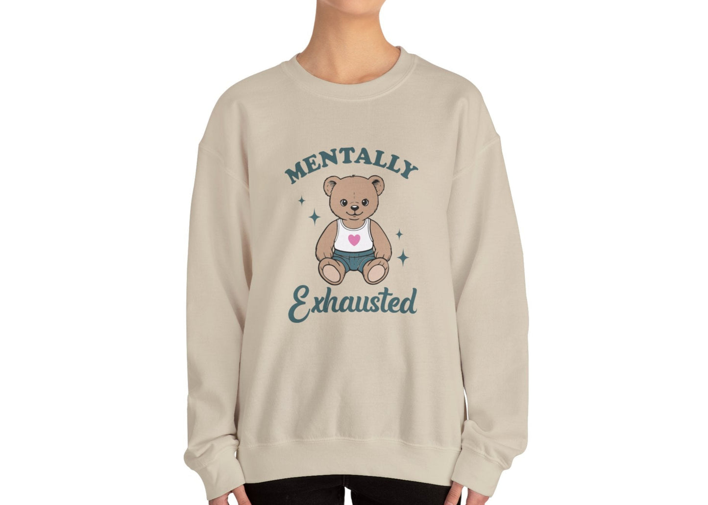 Model wearing a beige sweatshirt with a cute teddy bear wearing an apron and the text "Mentally Exhausted," standing with hands at sides. Part of the Vivid Divergence Sensory Friendly Unisex Sweater Jumper Range.