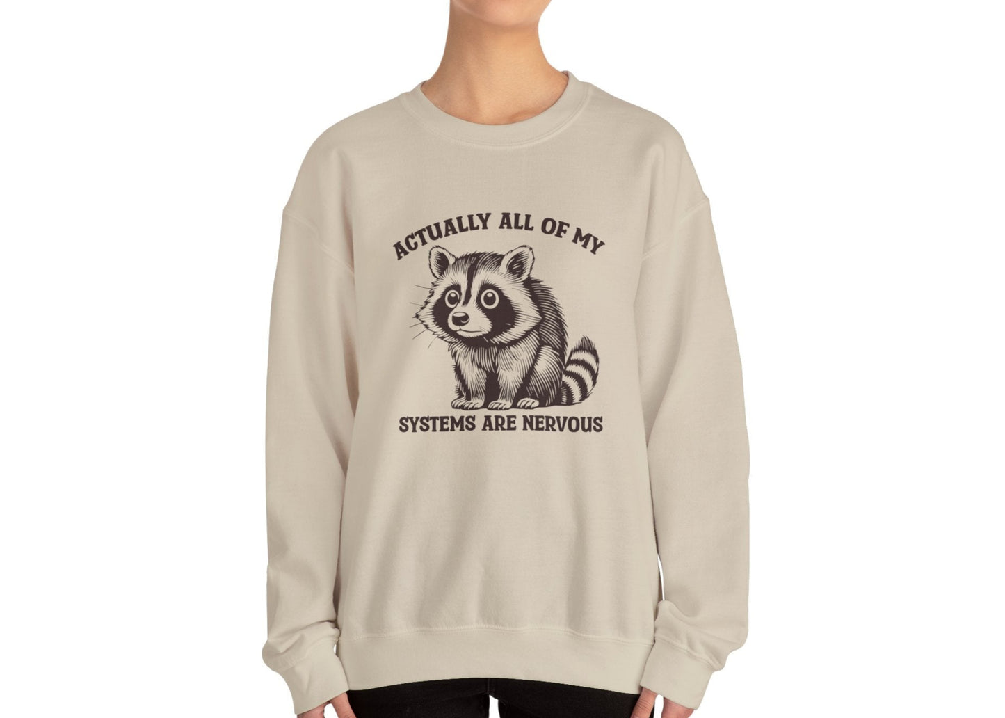 Model wearing a beige sweatshirt with a raccoon graphic and the text "Actually All of My Systems Are Nervous," standing with hands at sides. Part of the Vivid Divergence Sensory Friendly Unisex Sweater Jumper Range.