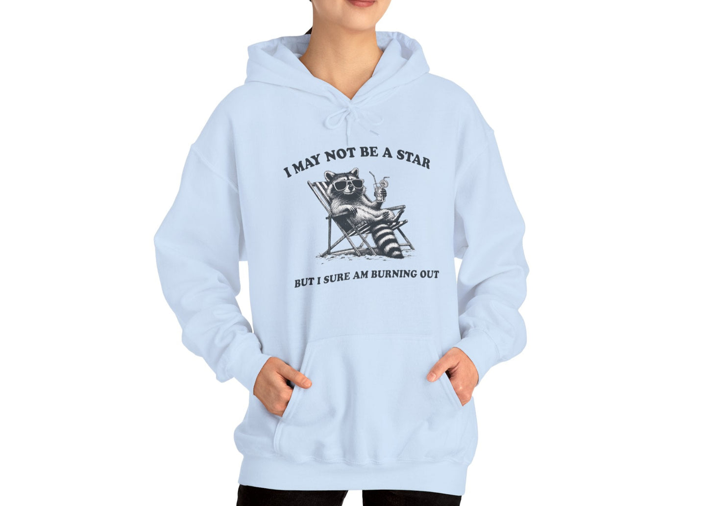 Model wearing a light blue hoodie with a raccoon sitting in a trash can and the text "I May Not Be A Star, But I Sure Am Burning Out," standing with hands in pockets. Part of the Vivid Divergence Sensory Friendly Unisex Hoodie Range