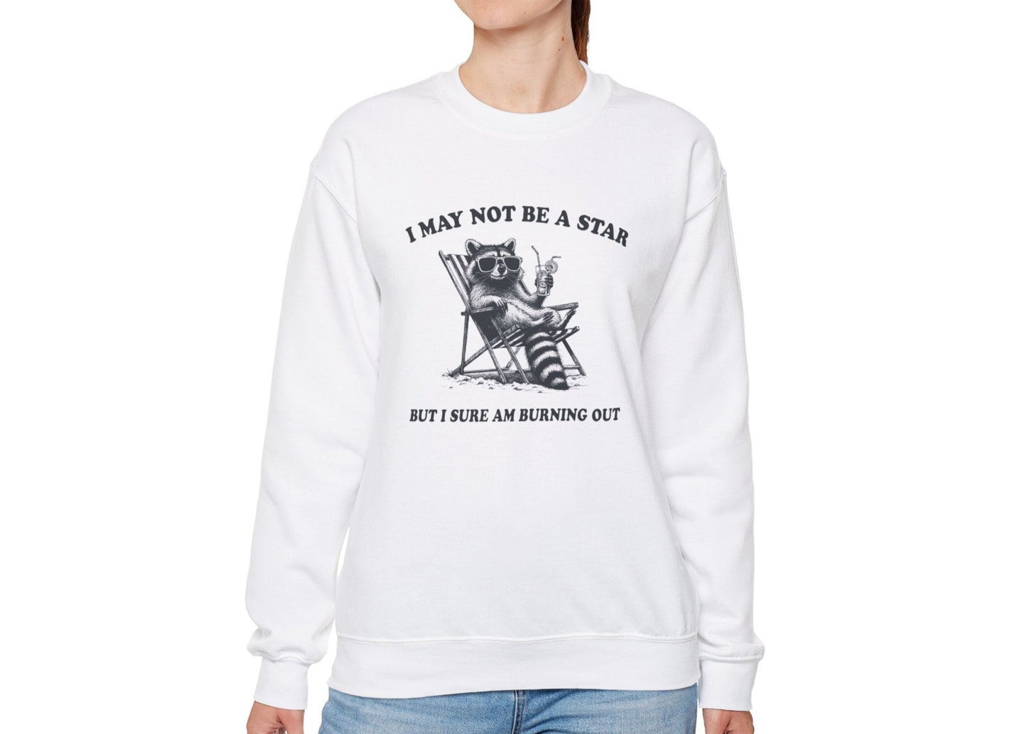 Model wearing a white sweatshirt with a raccoon sitting in a trash can and the text "I May Not Be A Star, But I Sure Am Burning Out," standing with hands at sides. Part of the Vivid Divergence Sensory Friendly Unisex Sweater Jumper Range.