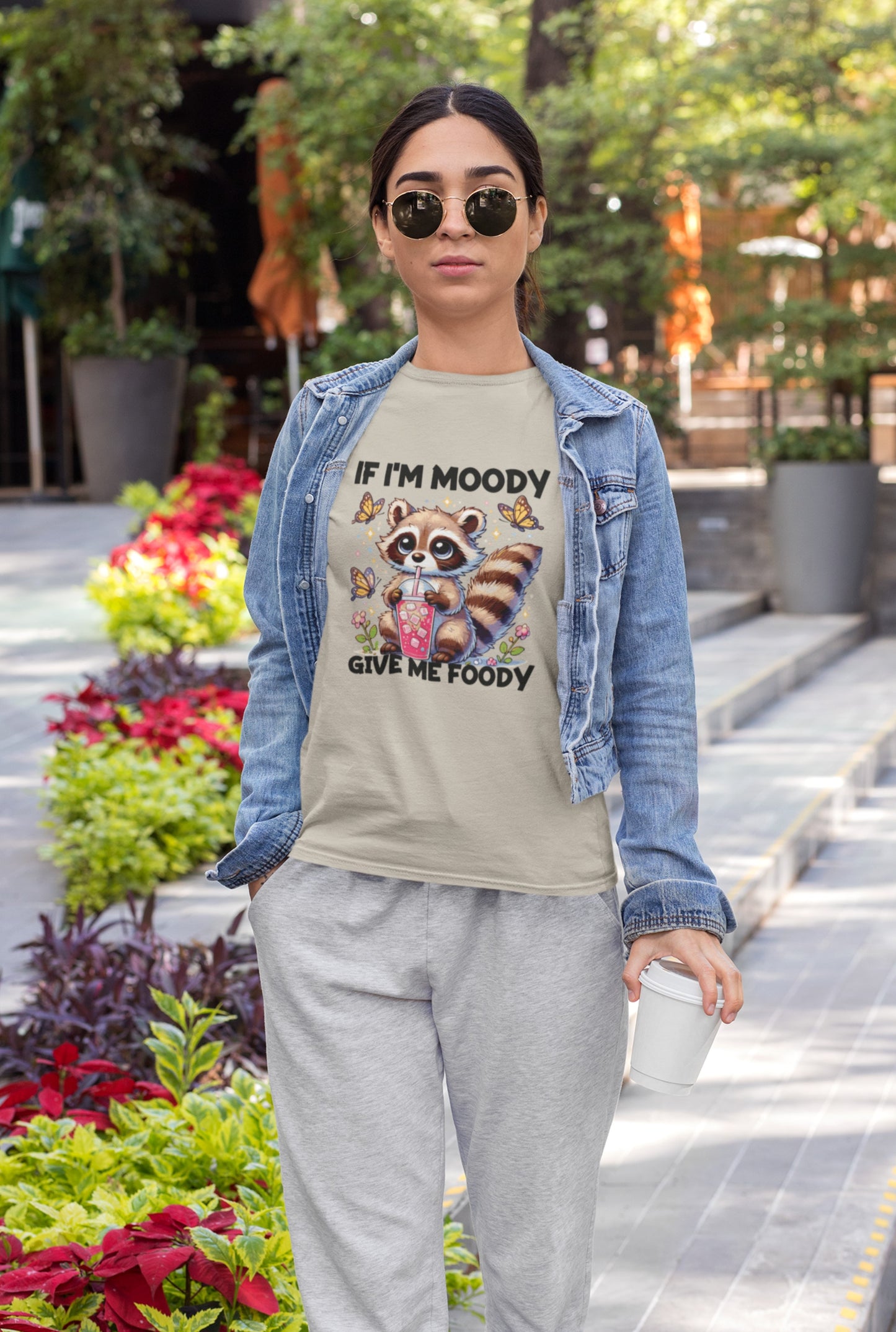 A person outdoors wearing a light blue T-shirt with a raccoon graphic and the text "If I'm Moody, Give Me Foody." Part of the Vivid Divergence Sensory Friendly Unisex Tees Range.