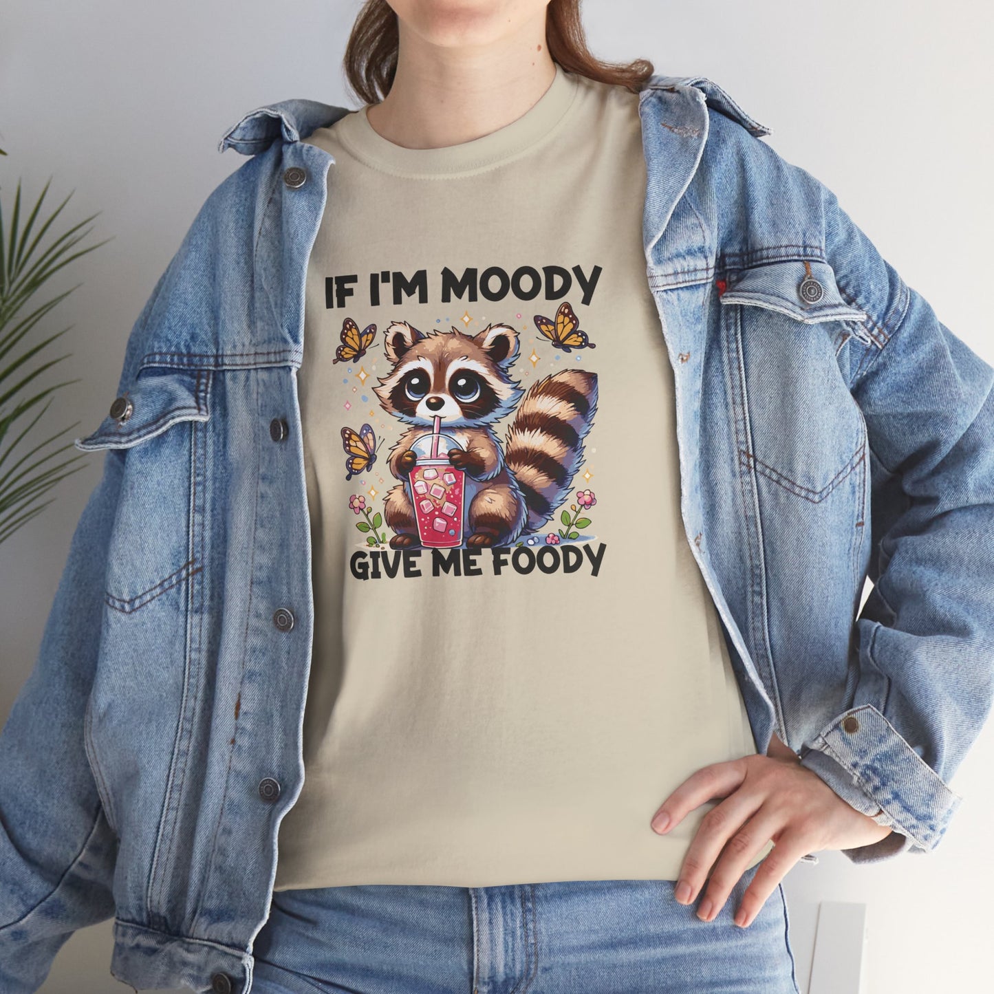 A person wearing a beige T-shirt with a raccoon graphic and the text "If I'm Moody, Give Me Foody." Part of the Vivid Divergence Sensory Friendly Unisex Tees Range.
