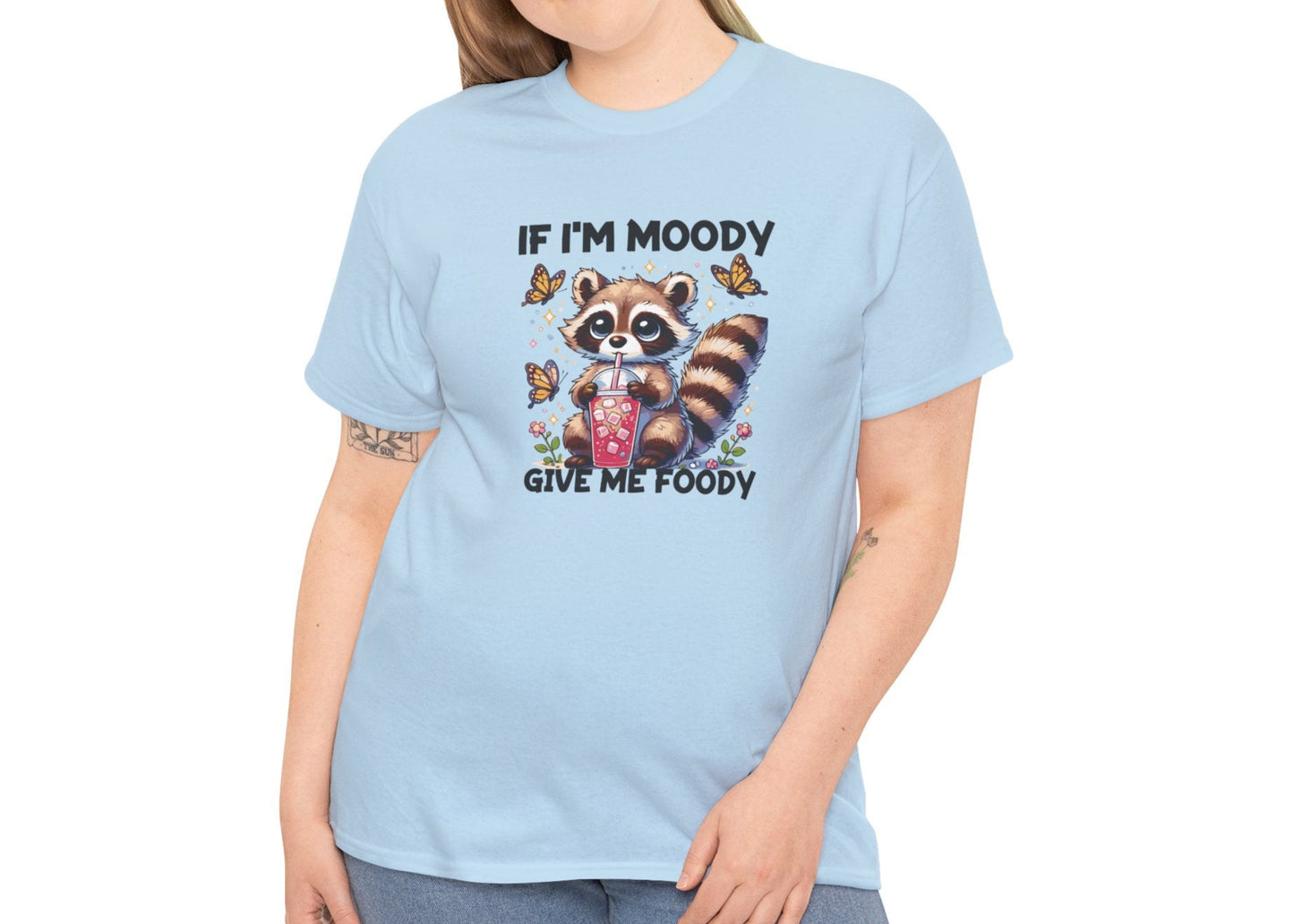 A person wearing a light blue T-shirt with a raccoon graphic and the text "If I'm Moody, Give Me Foody." Part of the Vivid Divergence Sensory Friendly Unisex Tees Range.