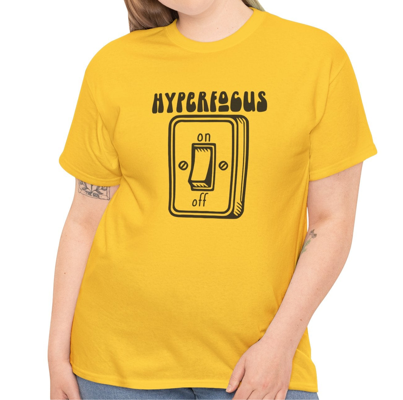 A person wearing a yellow T-shirt with the text "Hyperfocus" and an on/off switch graphic design. Part of the Vivid Divergence Sensory Friendly Unisex Tees Range.