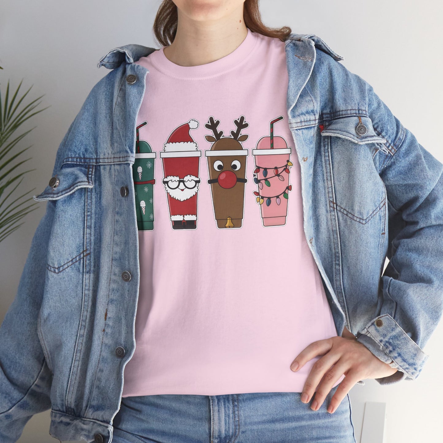 Model wearing a pink T-shirt featuring a festive illustration of four iced coffee cups decorated with Christmas designs, styled in a cozy indoor setting. Part of the Vivid Divergence Sensory Friendly Unisex Tees Range.