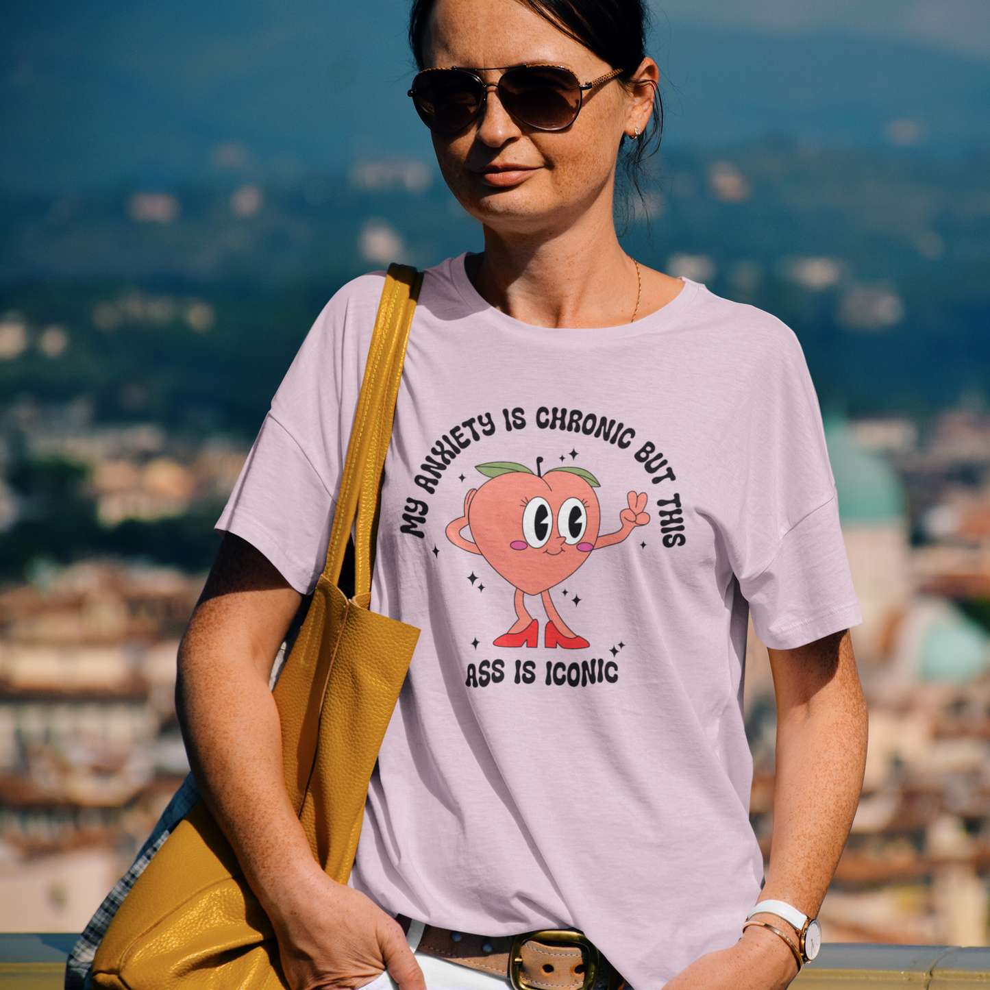 Woman wearing a pink T-shirt with the text "My Anxiety Is Chronic But This Ass Is Iconic" and a bold graphic, outdoors with sunglasses. Part of the Vivid Divergence Sensory Friendly Unisex Tees Range.