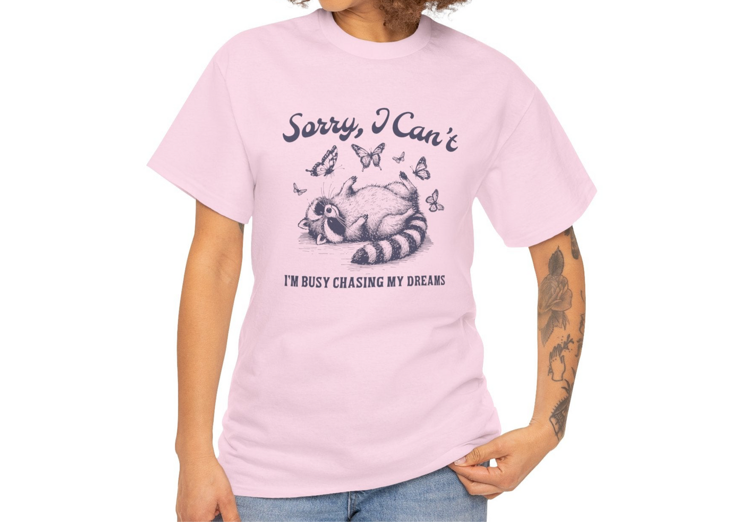 Model wearing a pink T-shirt with "Sorry I Can't, I'm Busy Chasing My Dreams" and a playful sleeping raccoon graphic in black text. Part of the Vivid Divergence Sensory Friendly Unisex Tees Range.