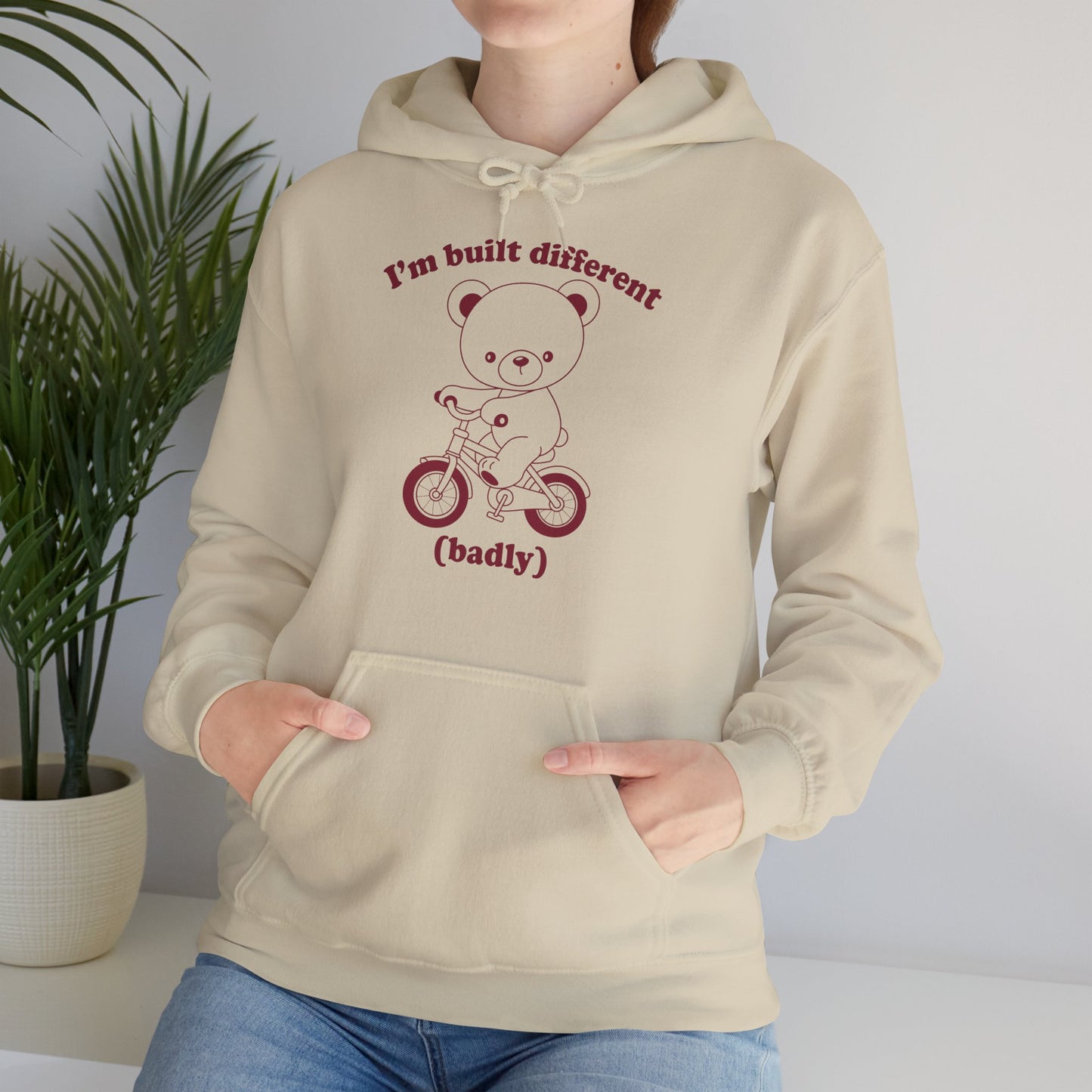 Model seated, wearing a beige hoodie with a cartoon bear riding a small bicycle and the text "I'm Built Different (Badly)," adjusting the hood. Part of the Vivid Divergence Sensory Friendly Unisex Hoodie Range.