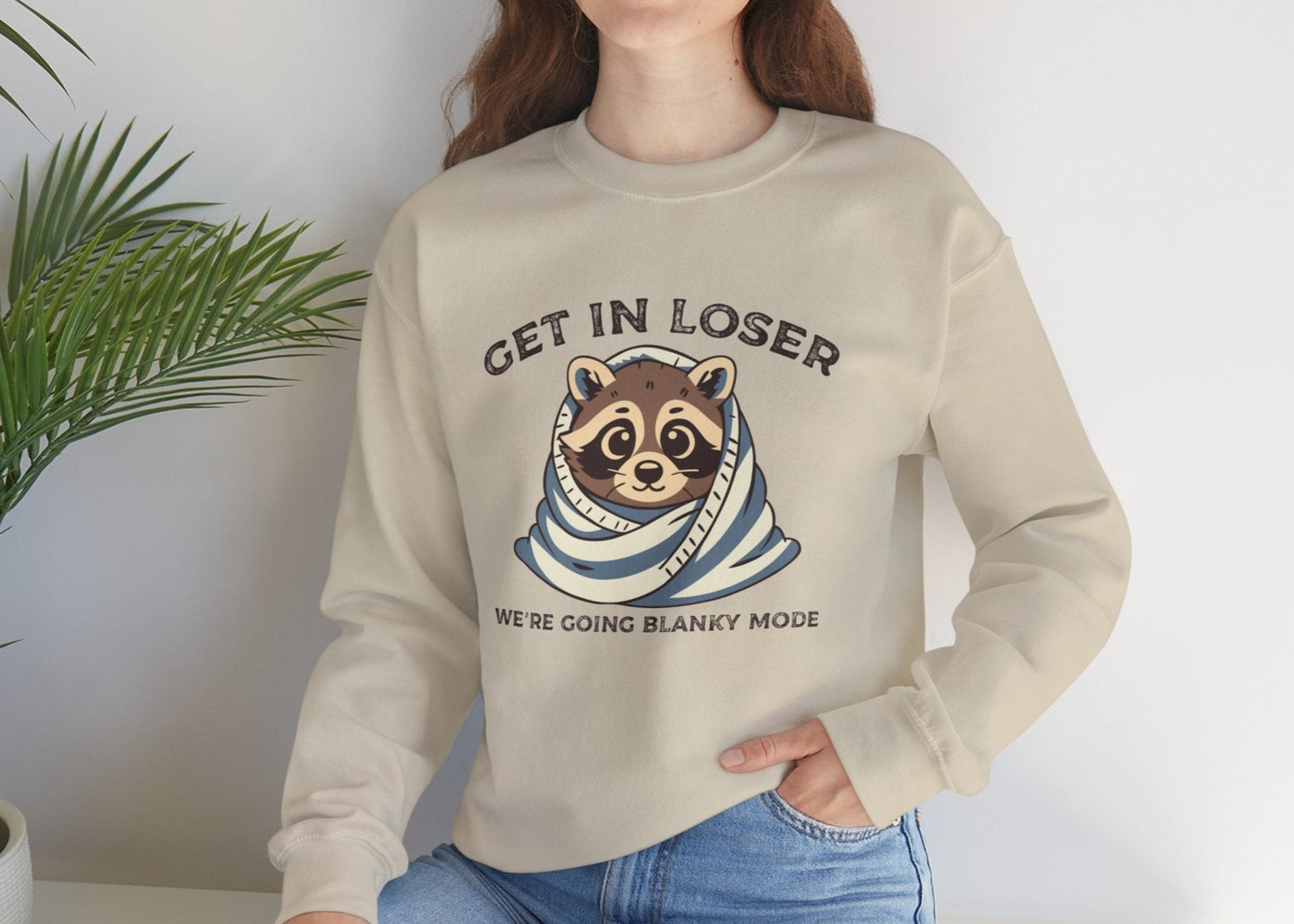 Model seated, wearing a beige sweatshirt with a raccoon wrapped in a blanket and the text "Get in Loser, We're Going Blanky Mode." Part of the Vivid Divergence Sensory Friendly Unisex Sweater Jumper Range.