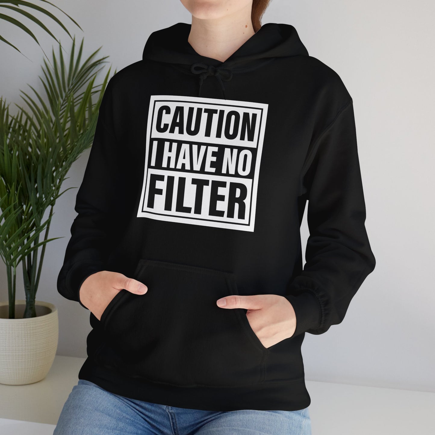 Model seated, wearing a black hoodie with bold white text inside a rectangular border that reads "Caution: I Have No Filter," adjusting the hood. Part of the Vivid Divergence Sensory Friendly Unisex Hoodie Range.