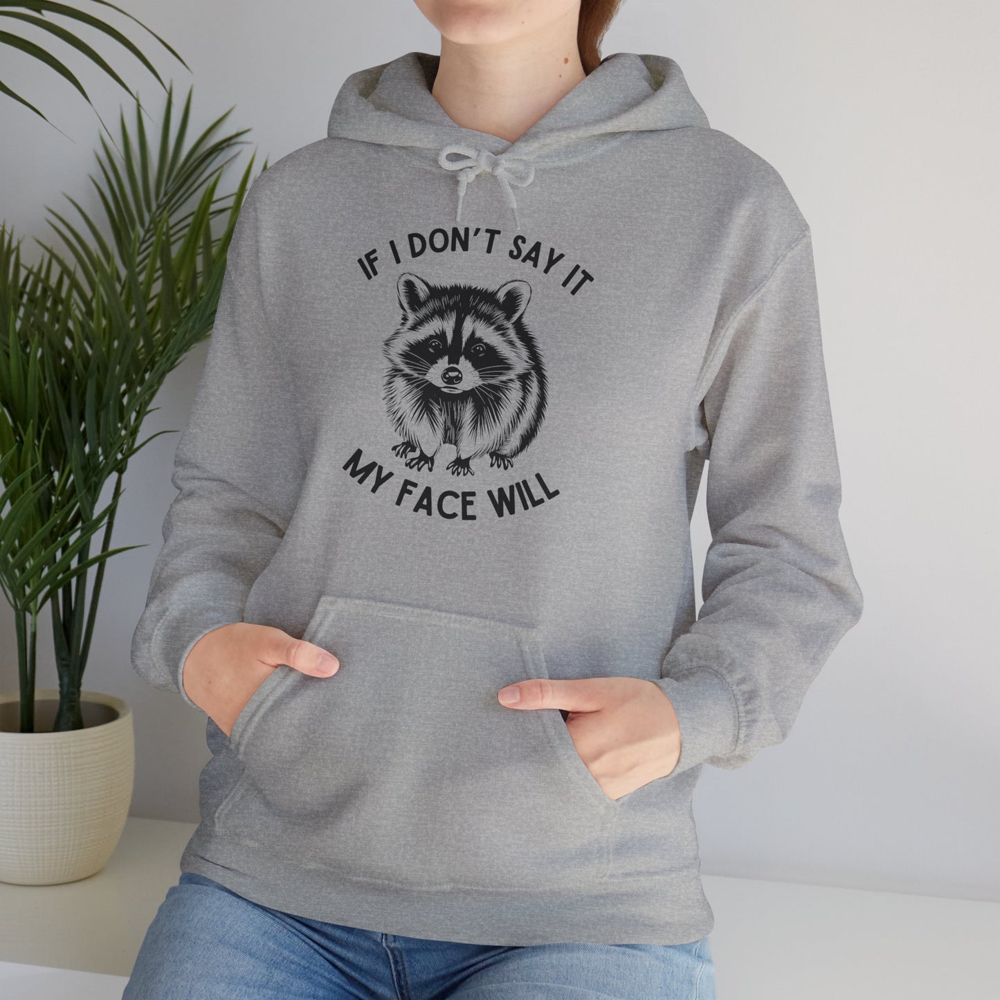 Model seated, wearing a gray hoodie with a raccoon graphic and the text "If I Don't Say It, My Face Will," adjusting the hood. Part of the Vivid Divergence Sensory Friendly Unisex Hoodie Range.