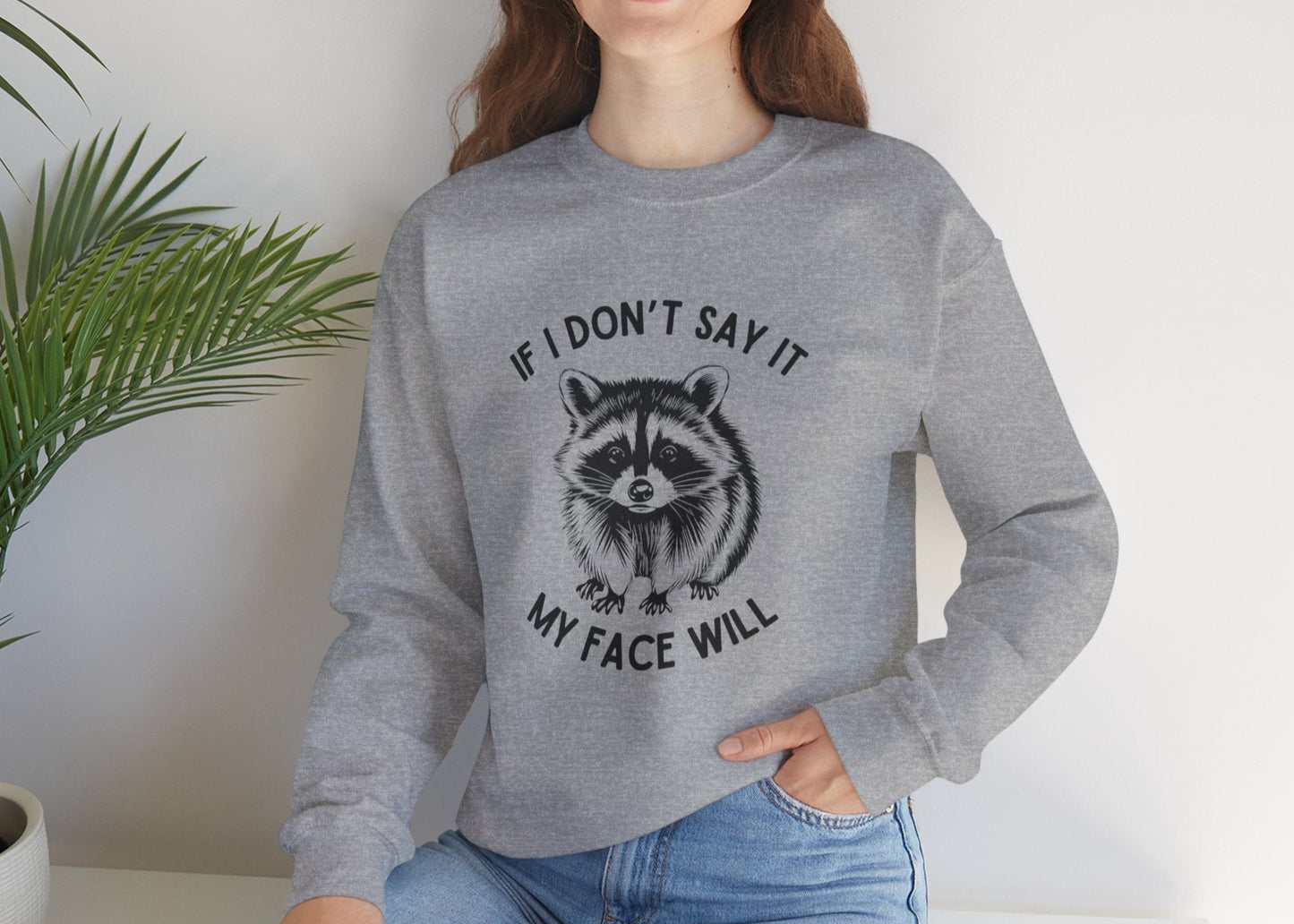 Model seated, wearing a gray sweatshirt with a raccoon graphic and the text "If I Don't Say It, My Face Will." Part of the Vivid Divergence Sensory Friendly Unisex Sweater Jumper Range.