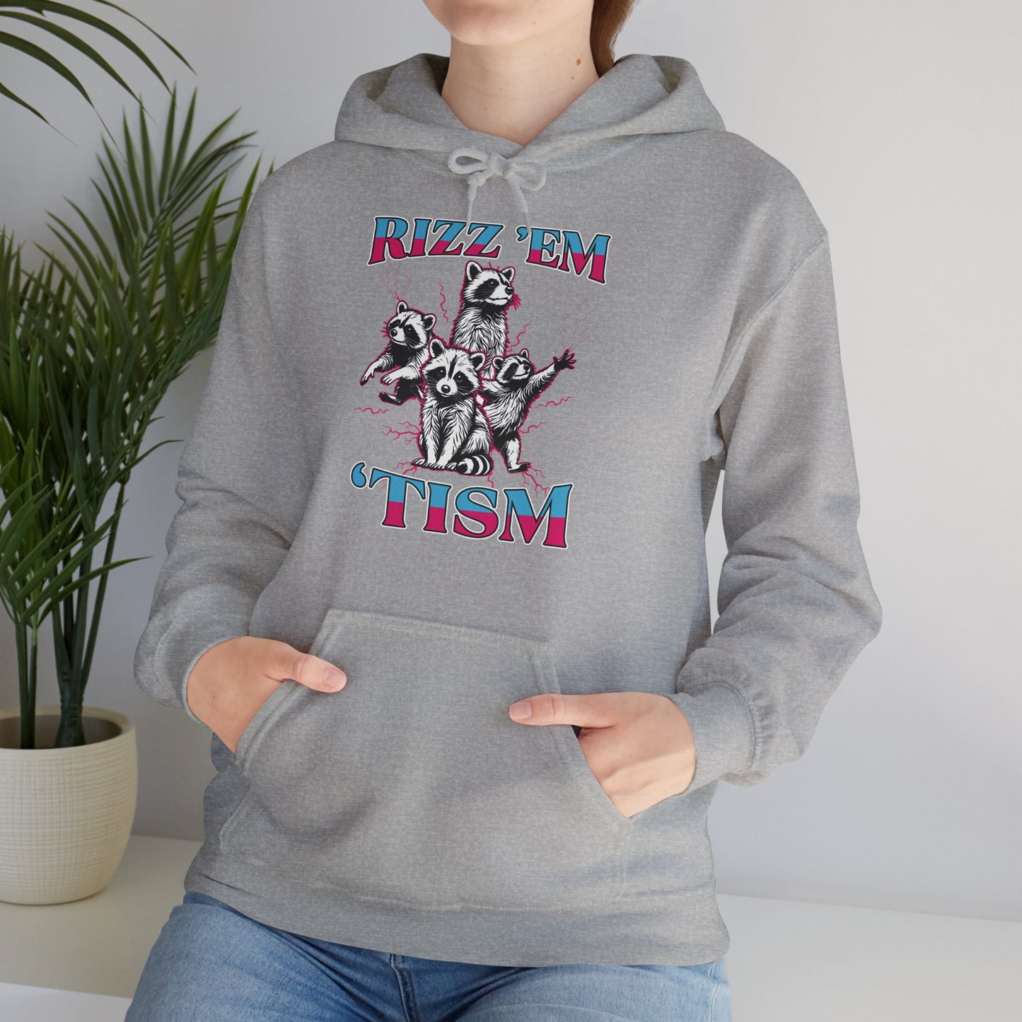 Model seated, wearing a gray hoodie with a raccoon illustration and the text "Rizz 'Em 'Tism," adjusting the hood. Part of the Vivid Divergence Sensory Friendly Unisex Hoodie Range.