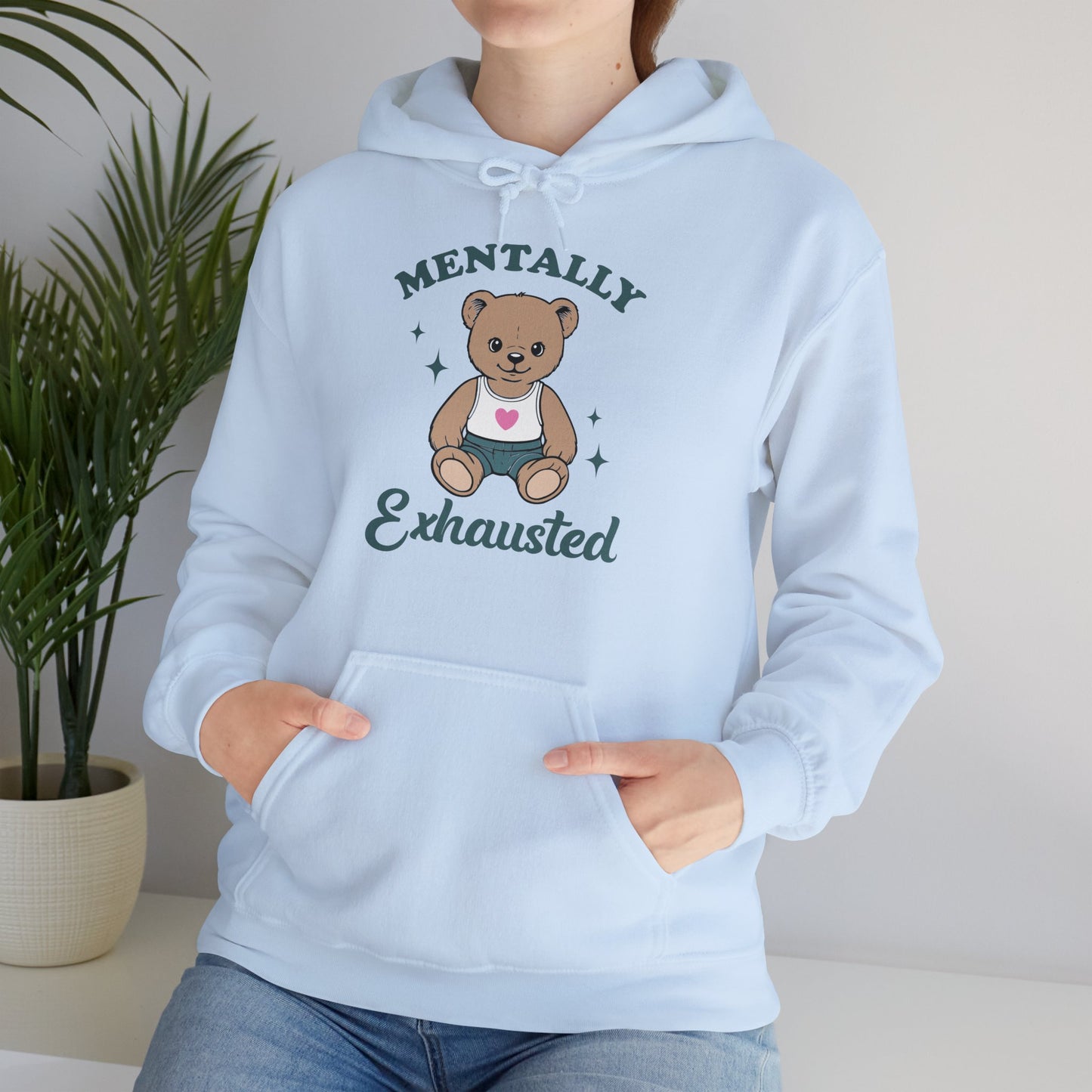 Model seated, wearing a light blue hoodie with a cute teddy bear wearing an apron and the text "Mentally Exhausted," adjusting the hood. Part of the Vivid Divergence Sensory Friendly Unisex Hoodie Range.