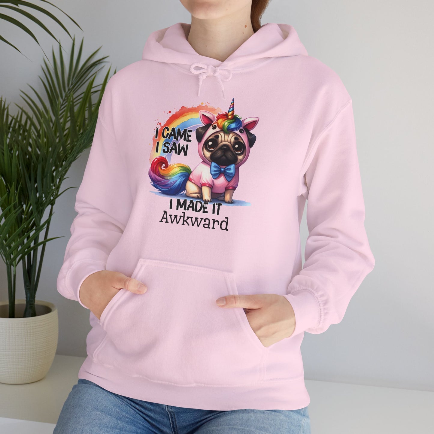 Model wearing a pink hoodie with a colorful unicorn pug and the text "I Came, I Saw, I Made It Awkward," seated with one hand in pocket. Part of the Vivid Divergence Sensory Friendly Unisex Hoodie Range.
