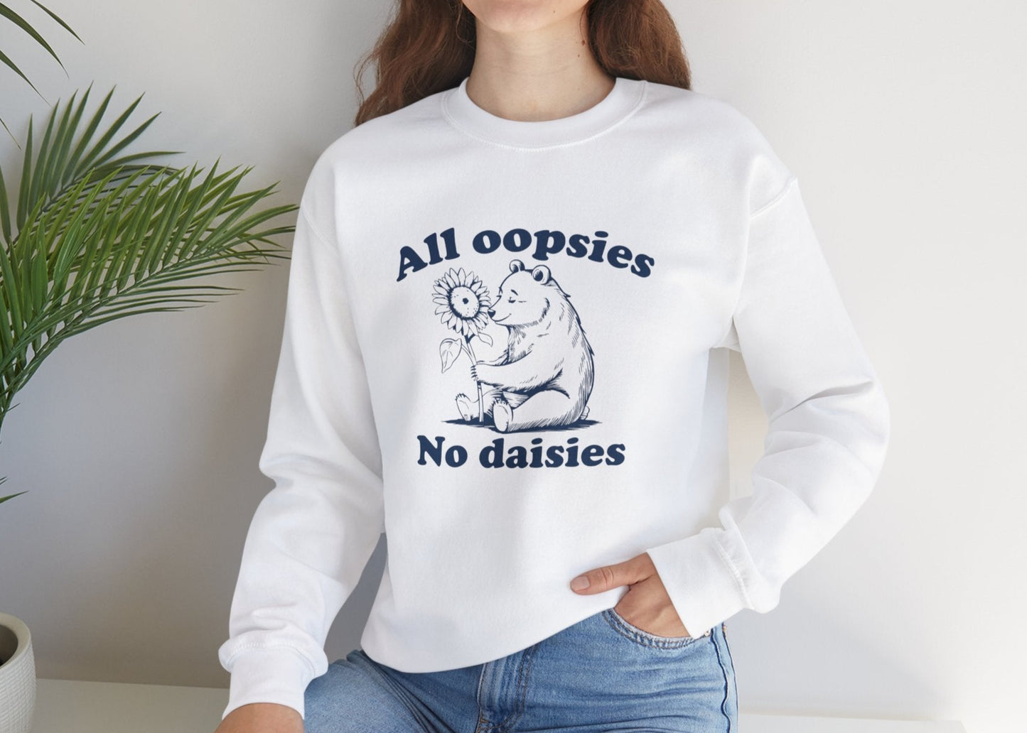 Model seated, wearing a white sweatshirt with a cartoon bear holding a sunflower and the text "All Oopsies, No Daisies." Part of the Vivid Divergence Sensory Friendly Unisex Sweater Jumper Range.
