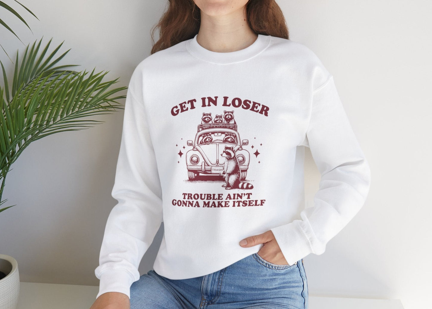 Model wearing a white sweatshirt with "Get in Loser, Trouble Ain't Gonna Cause Itself" in red letters and a group of raccoons in a car, seated with one hand on their knee. Part of the Vivid Divergence Sensory Friendly Unisex Sweater Jumper Range.