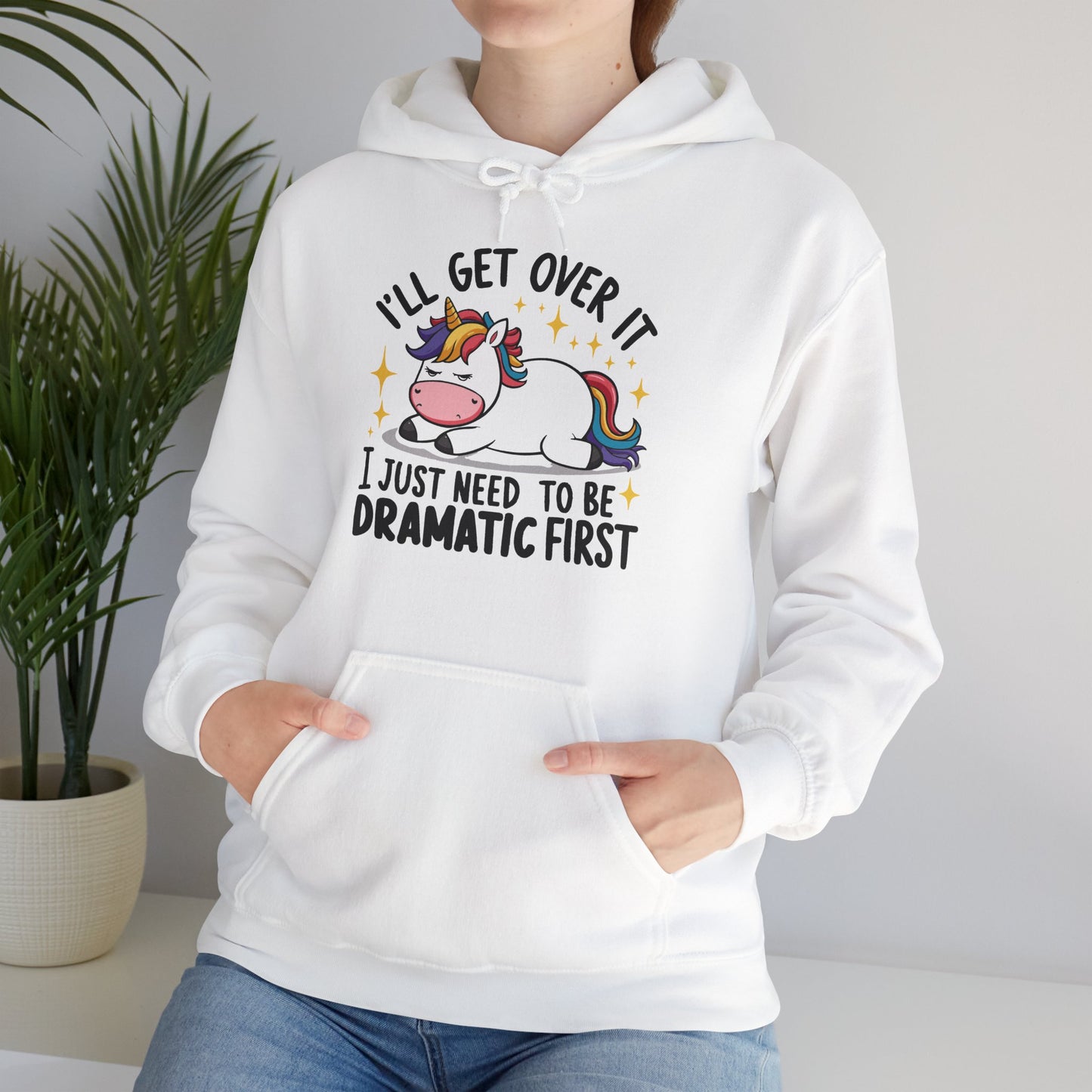 Model seated, wearing a white hoodie with a cartoon unicorn lying dramatically on a couch and the text "I'll Get Over It, I Just Need To Be Dramatic First," adjusting the hood. Part of the Vivid Divergence Sensory Friendly Unisex Hoodie Range.