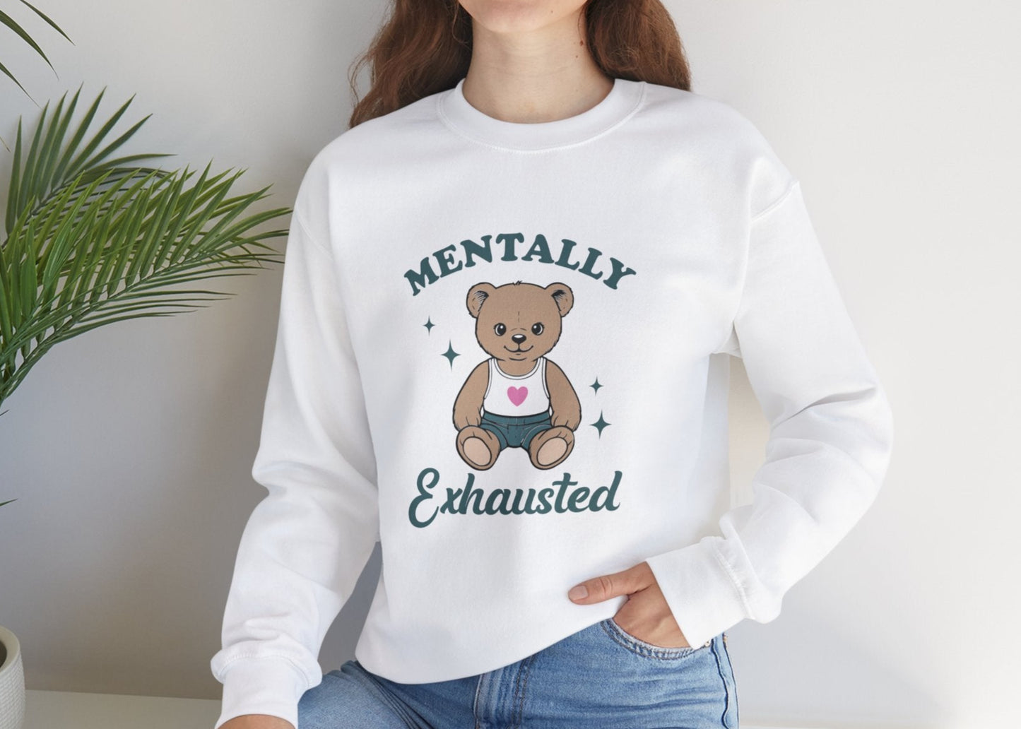 Model seated, wearing a white sweatshirt with a cute teddy bear wearing an apron and the text "Mentally Exhausted." Part of the Vivid Divergence Sensory Friendly Unisex Sweater Jumper Range.
