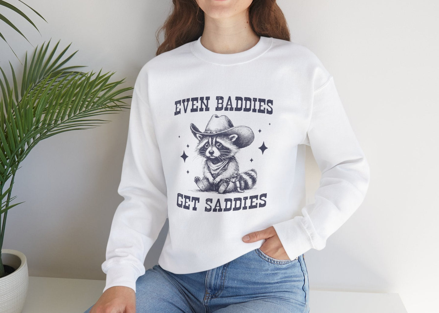 Model wearing a white sweater jumper with the text "Even Baddies Get Saddies" and a cartoon raccoon illustration. Part of the Vivid Divergence Sensory Friendly Unisex Sweater Jumper Range.