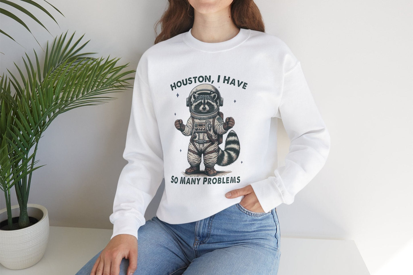 Model wearing a white sweater jumper with the text "Houston, I Have So Many Problems" and a cartoon raccoon illustration holding a striped object. Part of the Vivid Divergence Sensory Friendly Unisex Sweater Jumper Range.
