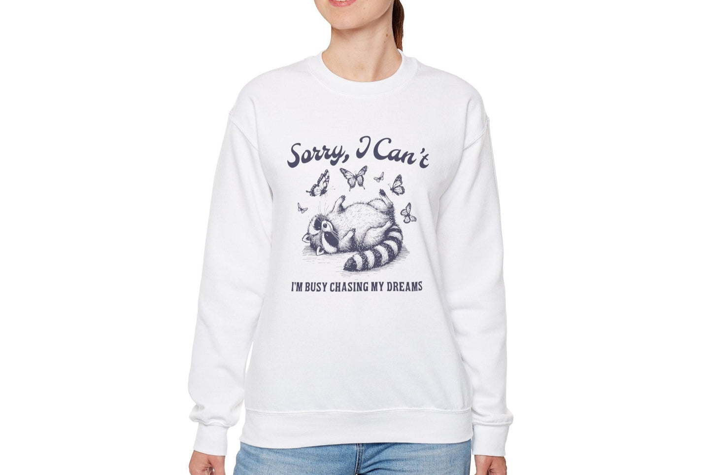 Model wearing a white sweater jumper with the text "Sorry, I Can't. I'm Busy Chasing My Dreams" and a cartoon raccoon illustration sleeping on a crescent moon. Part of the Vivid Divergence Sensory Friendly Unisex Sweater Jumper Range.