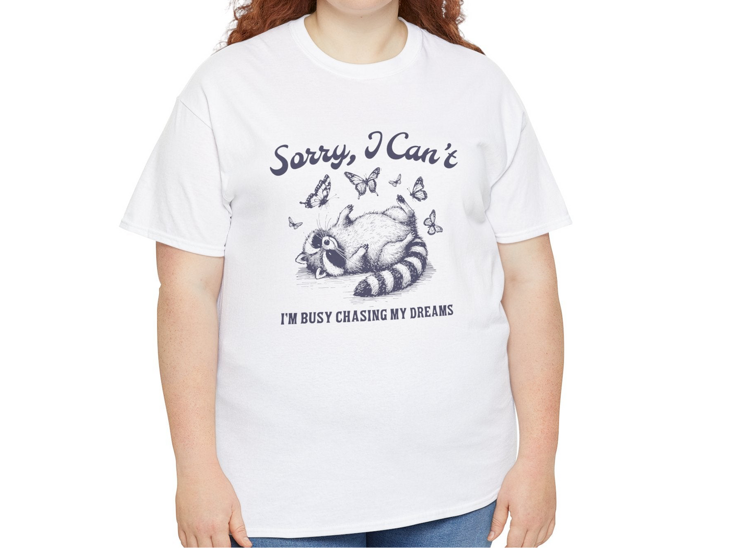 Model wearing a white T-shirt with "Sorry I Can't, I'm Busy Chasing My Dreams" and a playful sleeping raccoon graphic in black text. Part of the Vivid Divergence Sensory Friendly Unisex Tees Range.