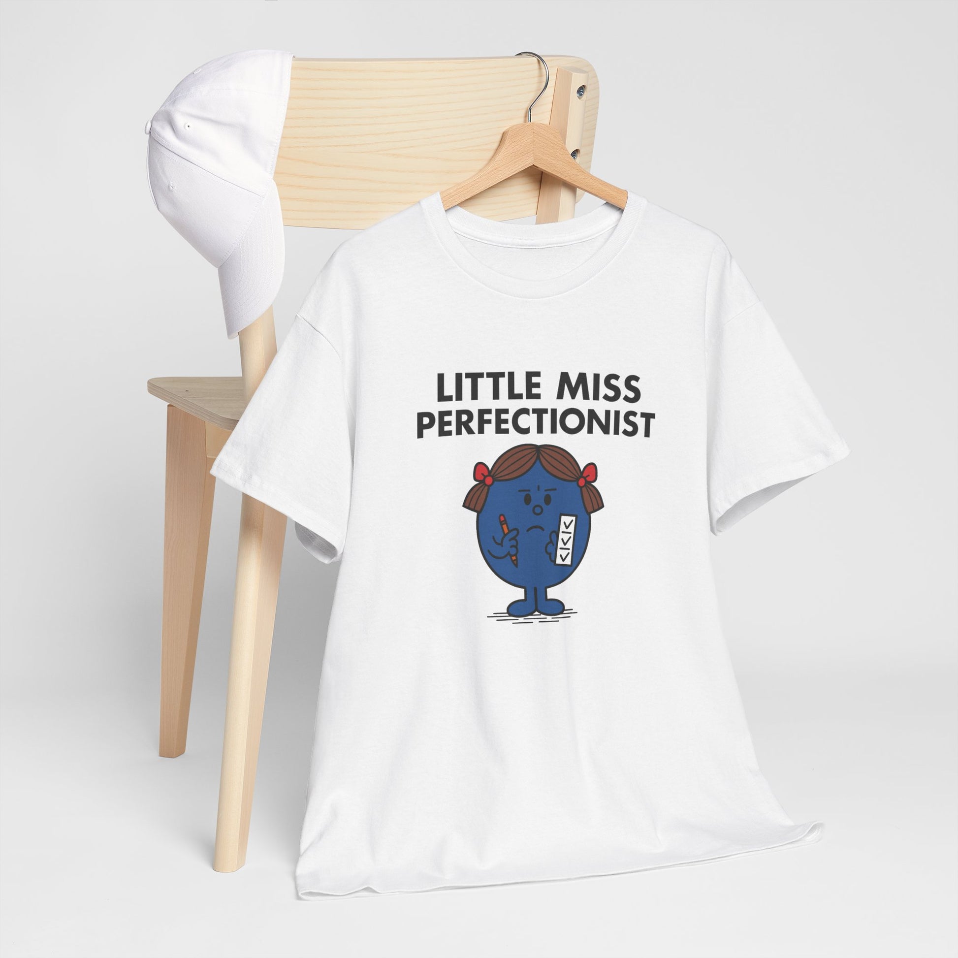 Little Miss Perfectionist T-shirt draped over a wooden chair in a minimal setup. Part of the Little Miss Meme Character Collection. A Vivid Divergence Sensory Friendly Unisex Tee.
