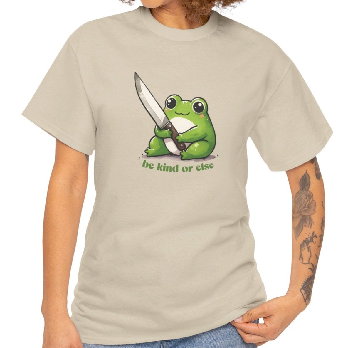 Beige T-shirt featuring a green frog holding a knife with the text "Be Kind or Else," modeled by a person standing. Part of the Vivid Divergence Sensory Friendly Unisex Tees Range.