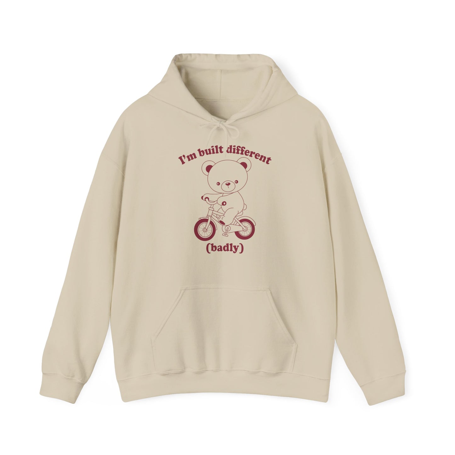 Front view of a beige hoodie featuring a cartoon bear riding a small bicycle with the text "I'm Built Different (Badly)." Part of the Vivid Divergence Sensory Friendly Unisex Hoodie Range.