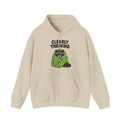 Front view of a beige hoodie featuring the text "Clearly Thriving" and a cartoon illustration of a raccoon wrapped in a cozy blanket. Part of the Vivid Divergence Sensory Friendly Unisex Hoodie Range.